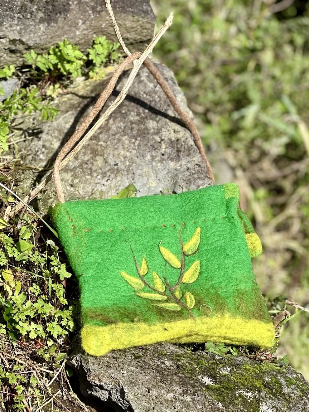 A small, felted bag with a bright green and yellow leaf design, resting on a rock outdoors surrounded by small green plants.