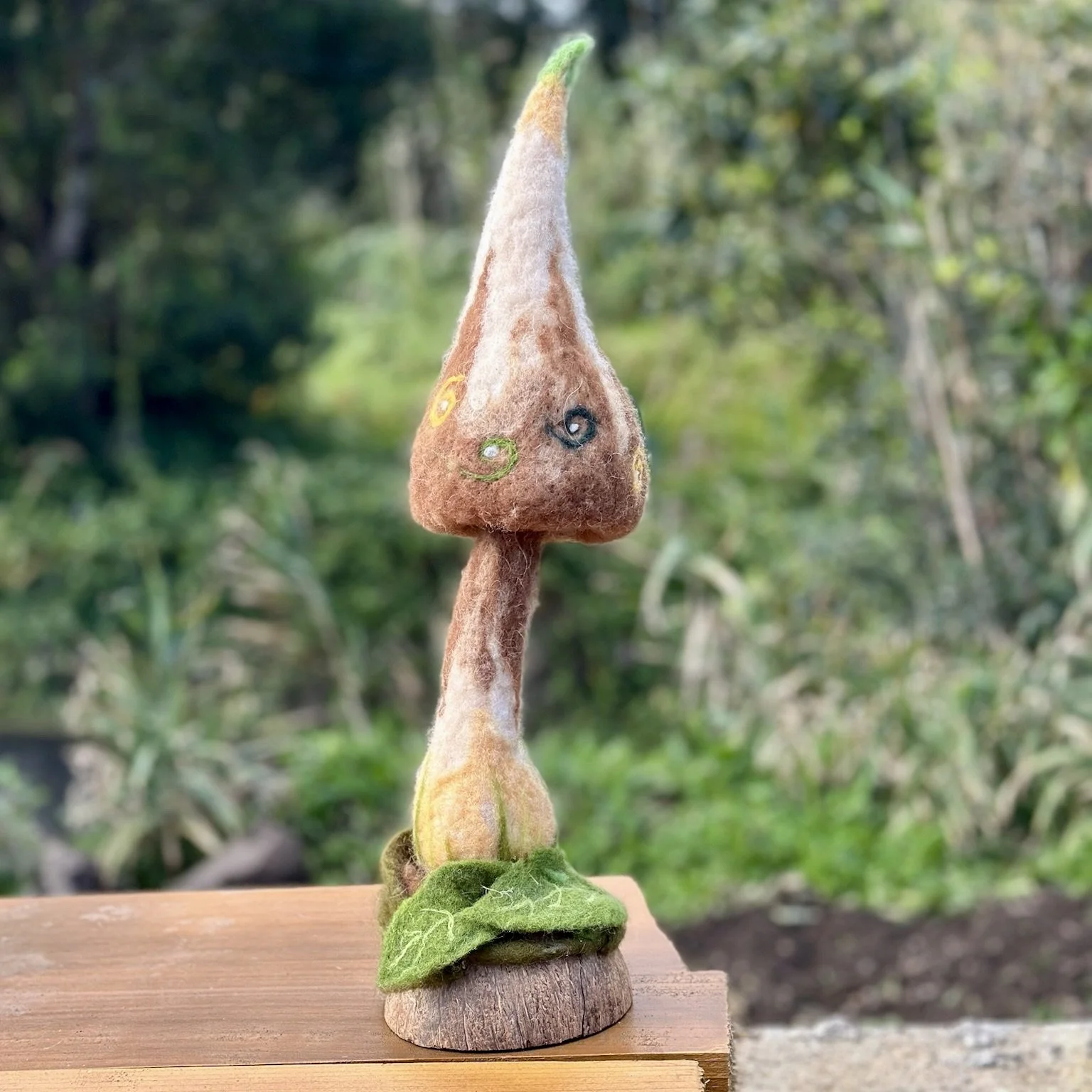 Felted mushroom sculpture with a brown and white cap, green details, and a green leaf at the base, placed on a wooden surface outdoors.
