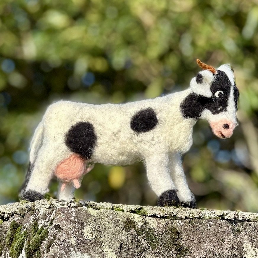 Felted toy cow with black and white spots, pink udder, and small horns on a tree stump.
