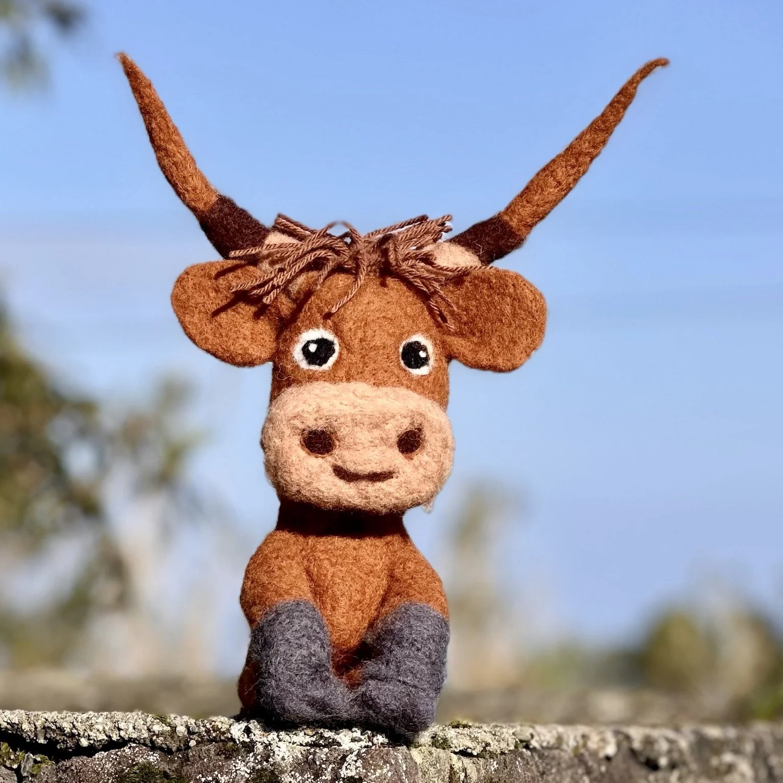 Felt plush toy of a brown and gray deer with big eyes, sitting outdoors on a branch with a clear blue sky in the background.
