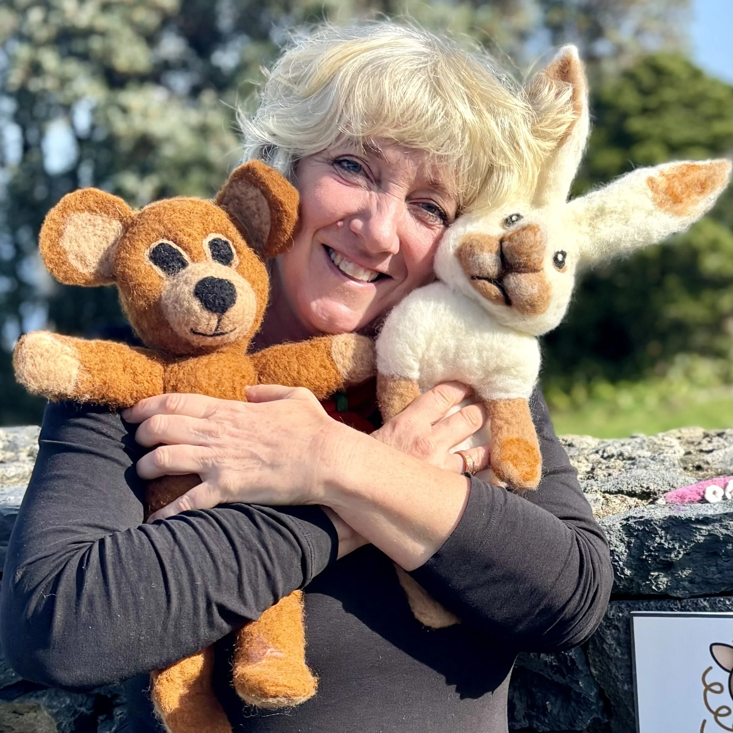 A woman is smiling and hugging two handmade stuffed animals, a bear and a rabbit, outdoors with trees in the background.