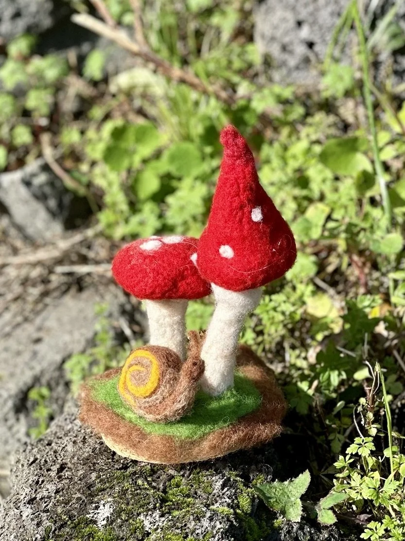 Felted mushroom and snail figurine on a piece of felted land, outdoors among greenery.