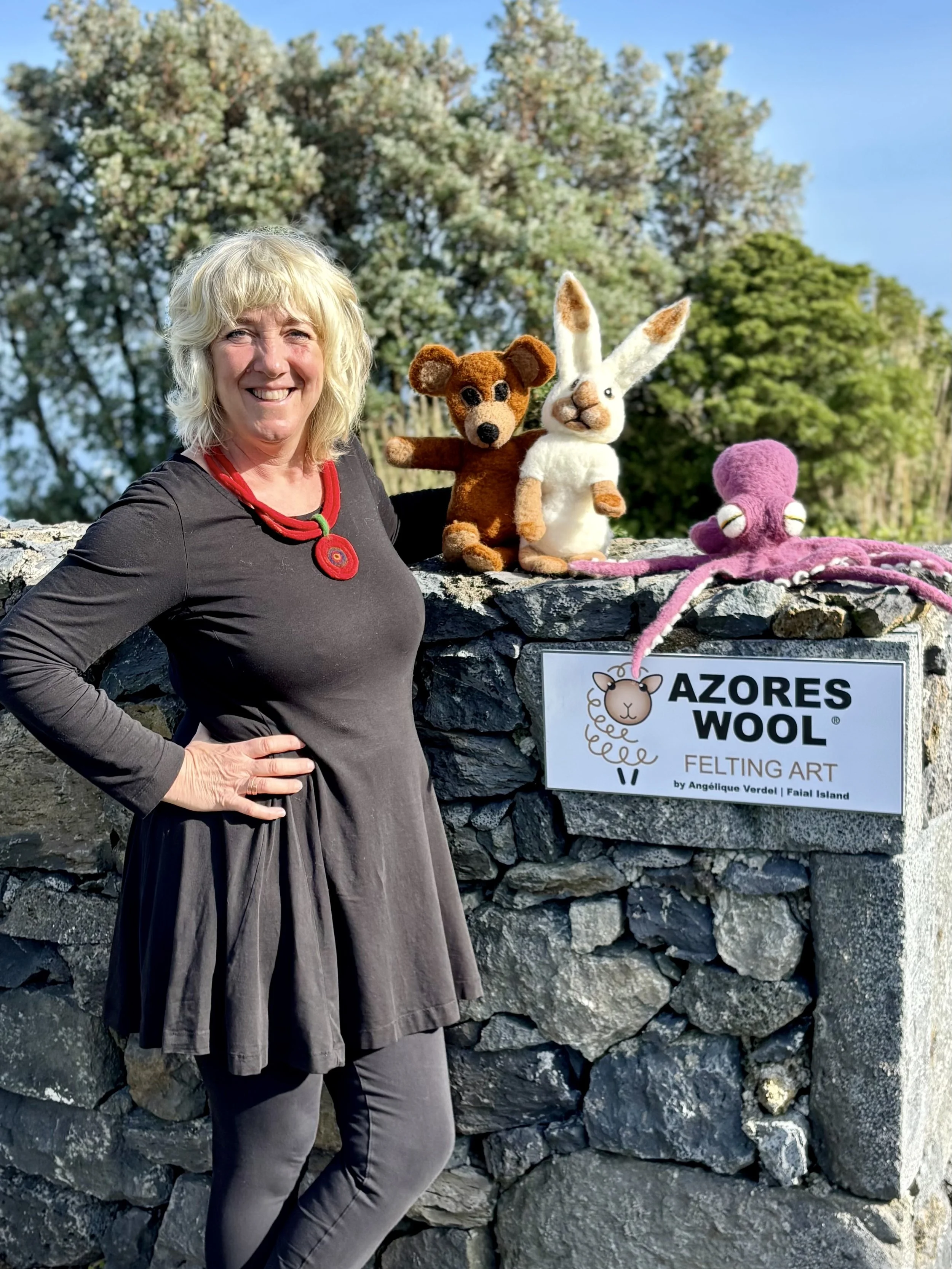 Woman in black dress standing next to a stone wall with plush felted animal figures and a sign that reads 'Azores Wool Felting Art by Angélique Verdel, Faial Island'.