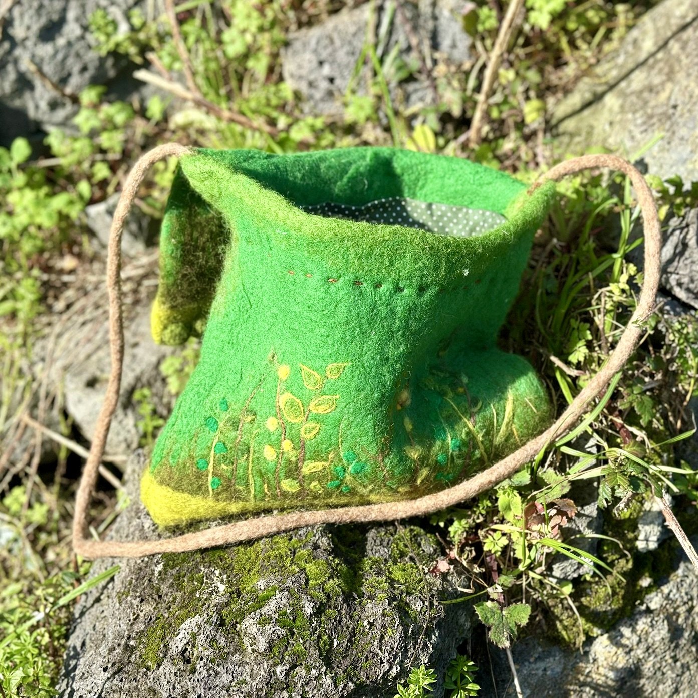 A small, bright green felt pouch with embroidered yellow and green details, resting on a moss-covered rock surrounded by small plants and grass.