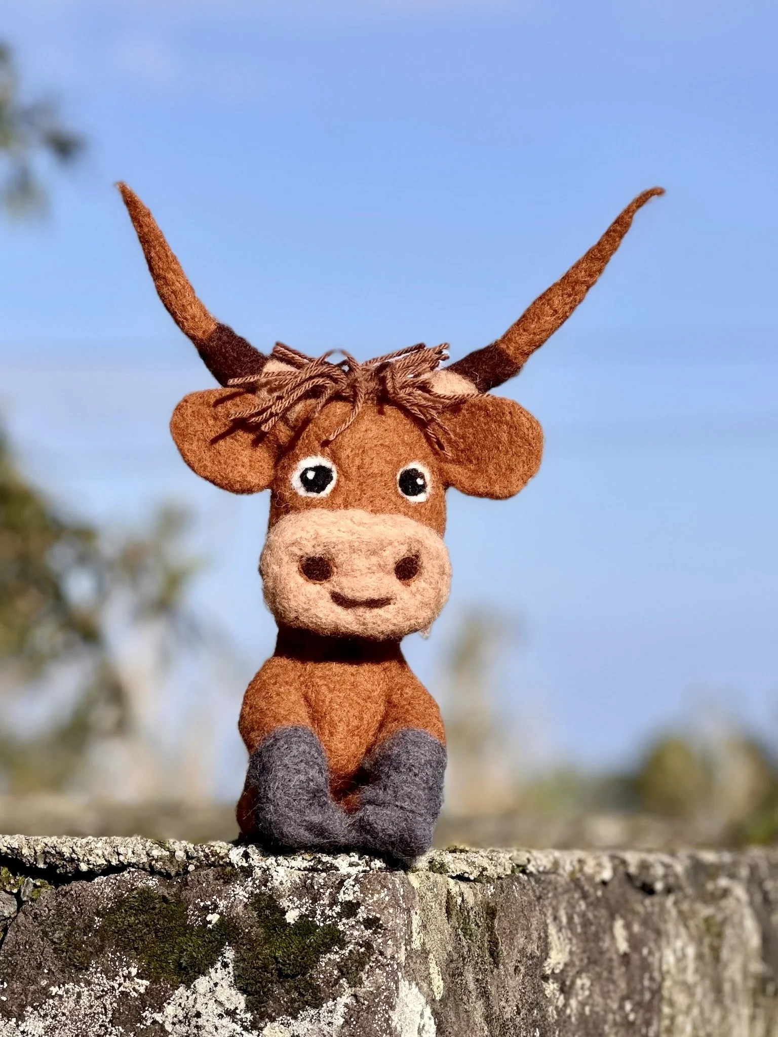 A small plush toy of a cartoon-style deer with large eyes, brown body, and dark gray feet, sitting on a stone wall with a blurred outdoor background and a blue sky.