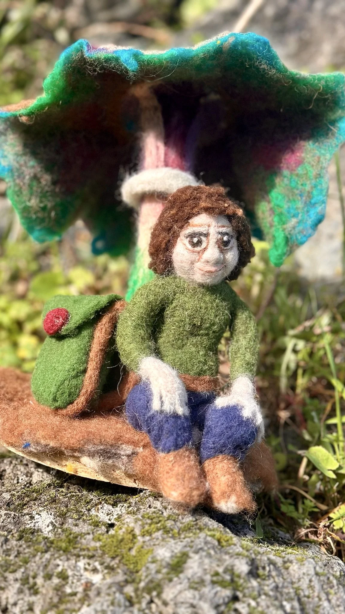 Felted doll of a woman sitting outdoors on a rock, with a backpack and a colorful umbrella in the background.