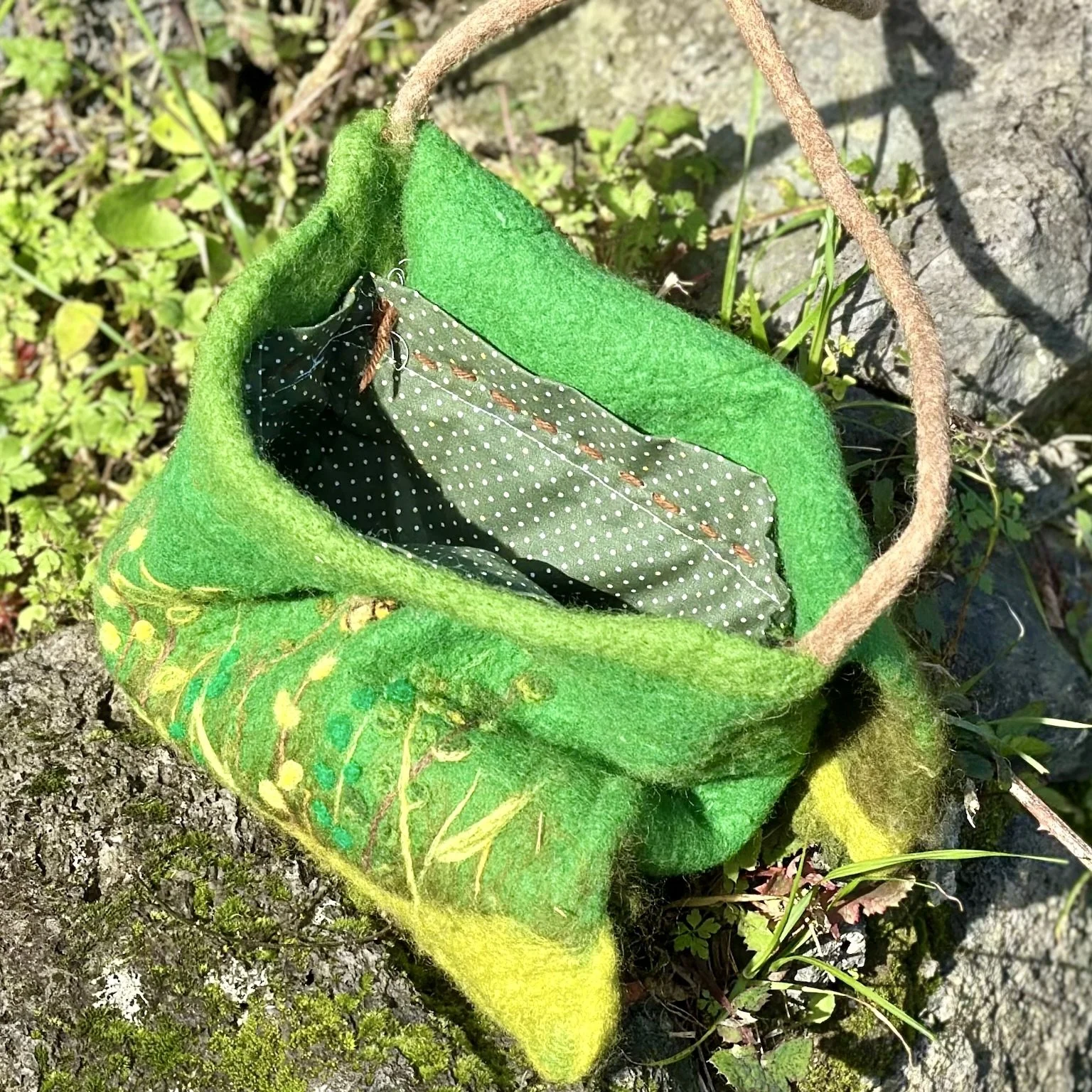 A green felt bag with a brown handle, opened to show a black interior with white polka dots, resting on a tree branch with moss and small plants nearby.
