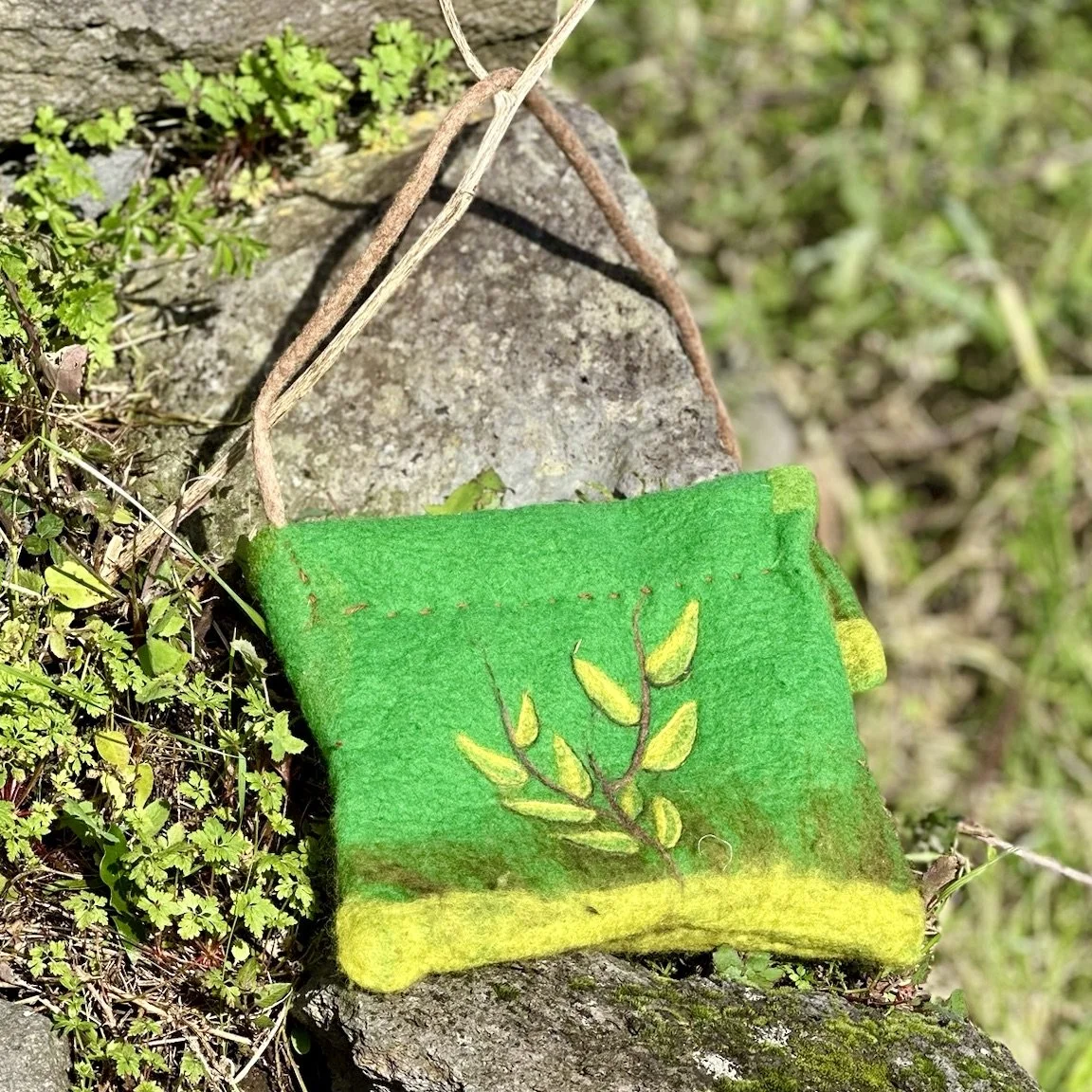A small, colorful felt pouch with a green and yellow design hanging from a branch outdoors.