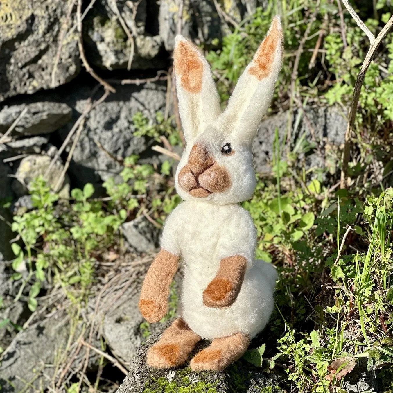 A handmade felted bunny toy with large ears, sitting outdoors on a piece of wood among grass and small plants.