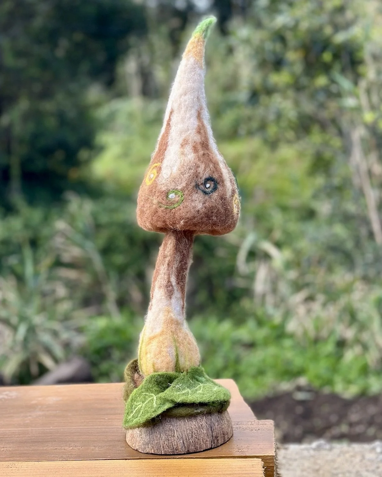 A handmade felted mushroom sculpture with a brown cap decorated with colorful spirals, a white and brown stem, a green felted leaf at the base, and a round wooden platform, set outdoors with a lush green background.