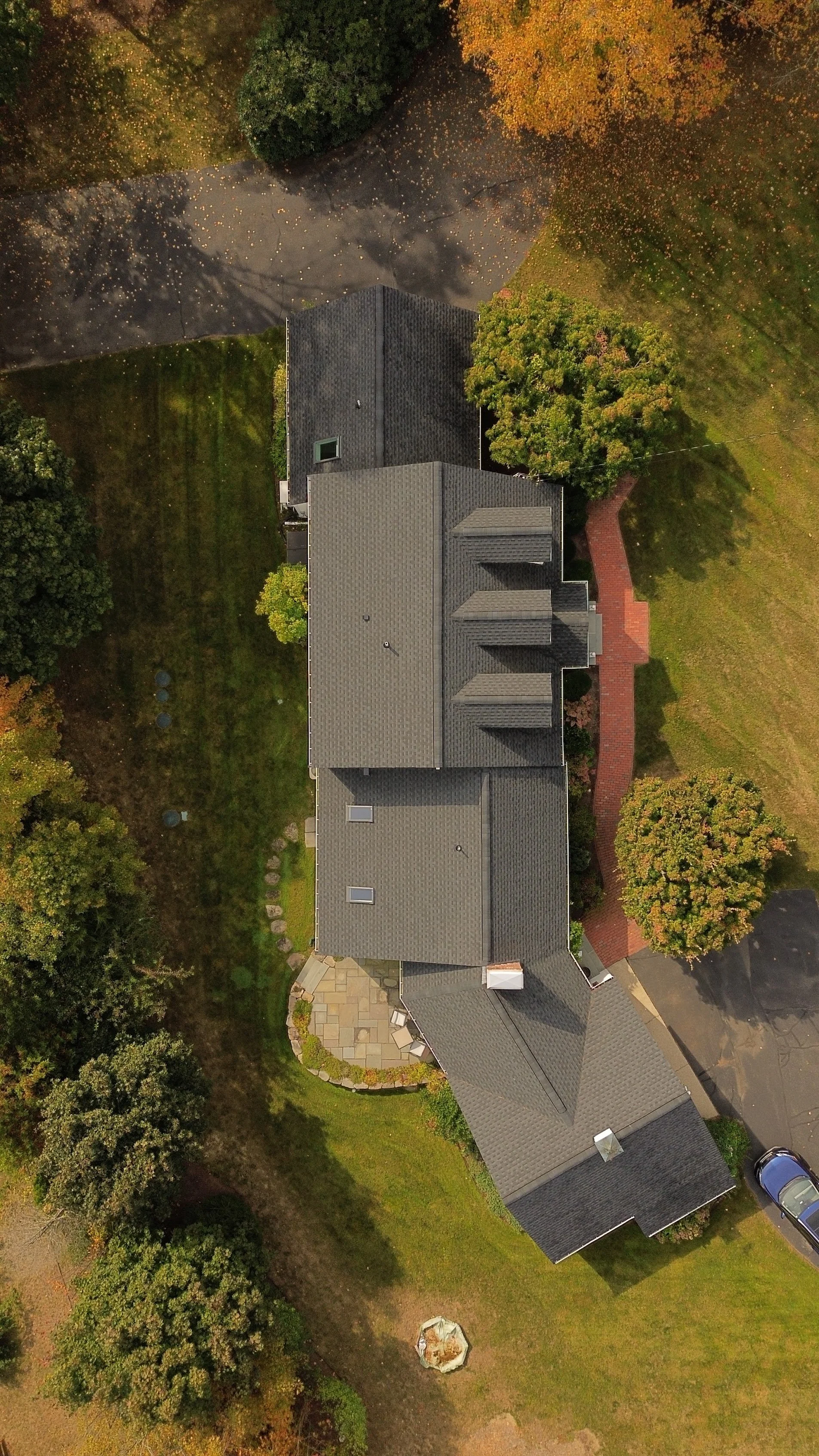 An aerial view of a house with a dark gray roof surrounded by trees with green and autumn-colored leaves, a curved brick walkway leading to the side entrance, and a paved driveway with parked cars.