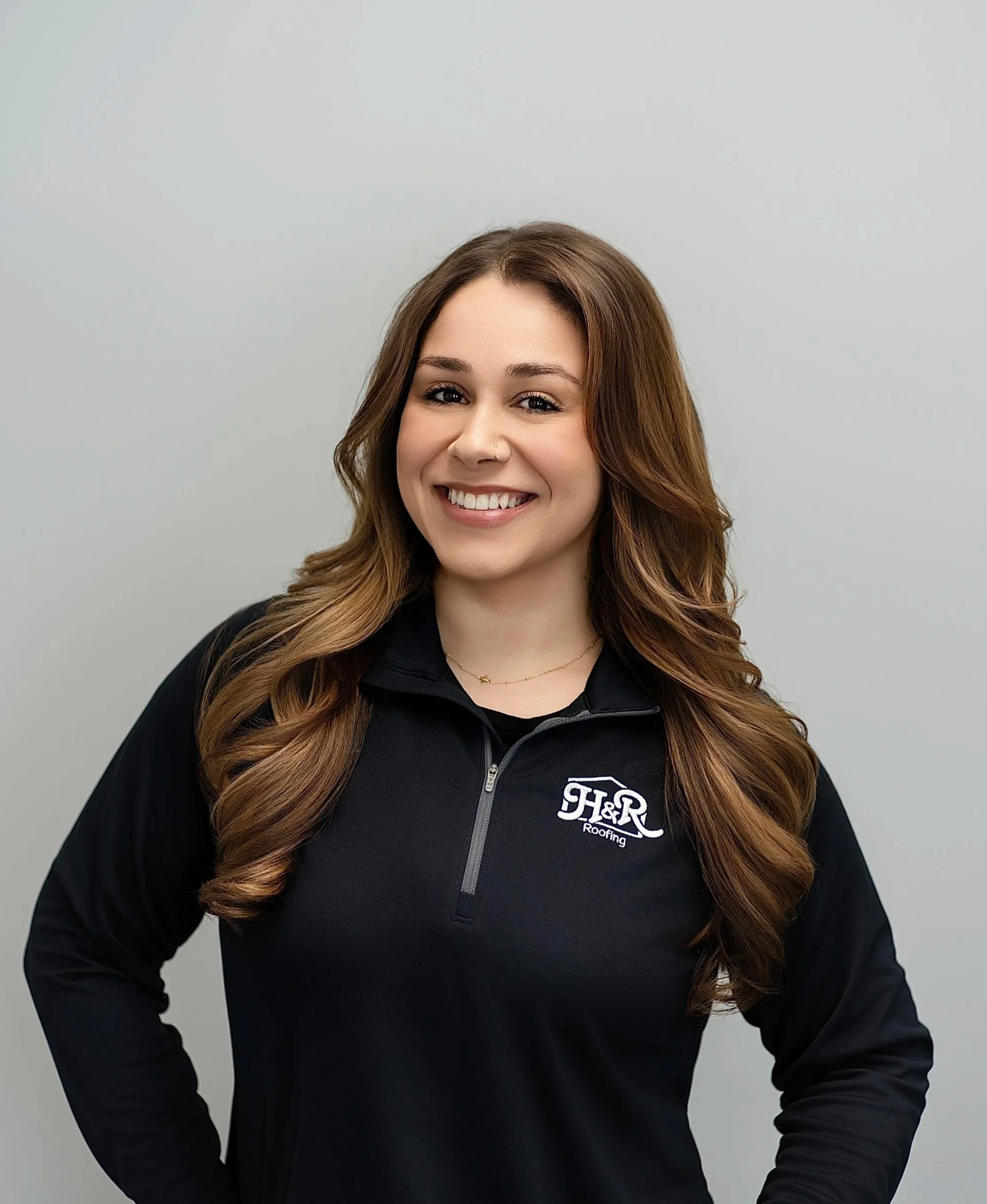 Young woman with long, wavy brown hair smiling, wearing a black quarter-zip jacket with a logo that says 'H&R Roofing' on the left chest, standing against a plain gray background.
