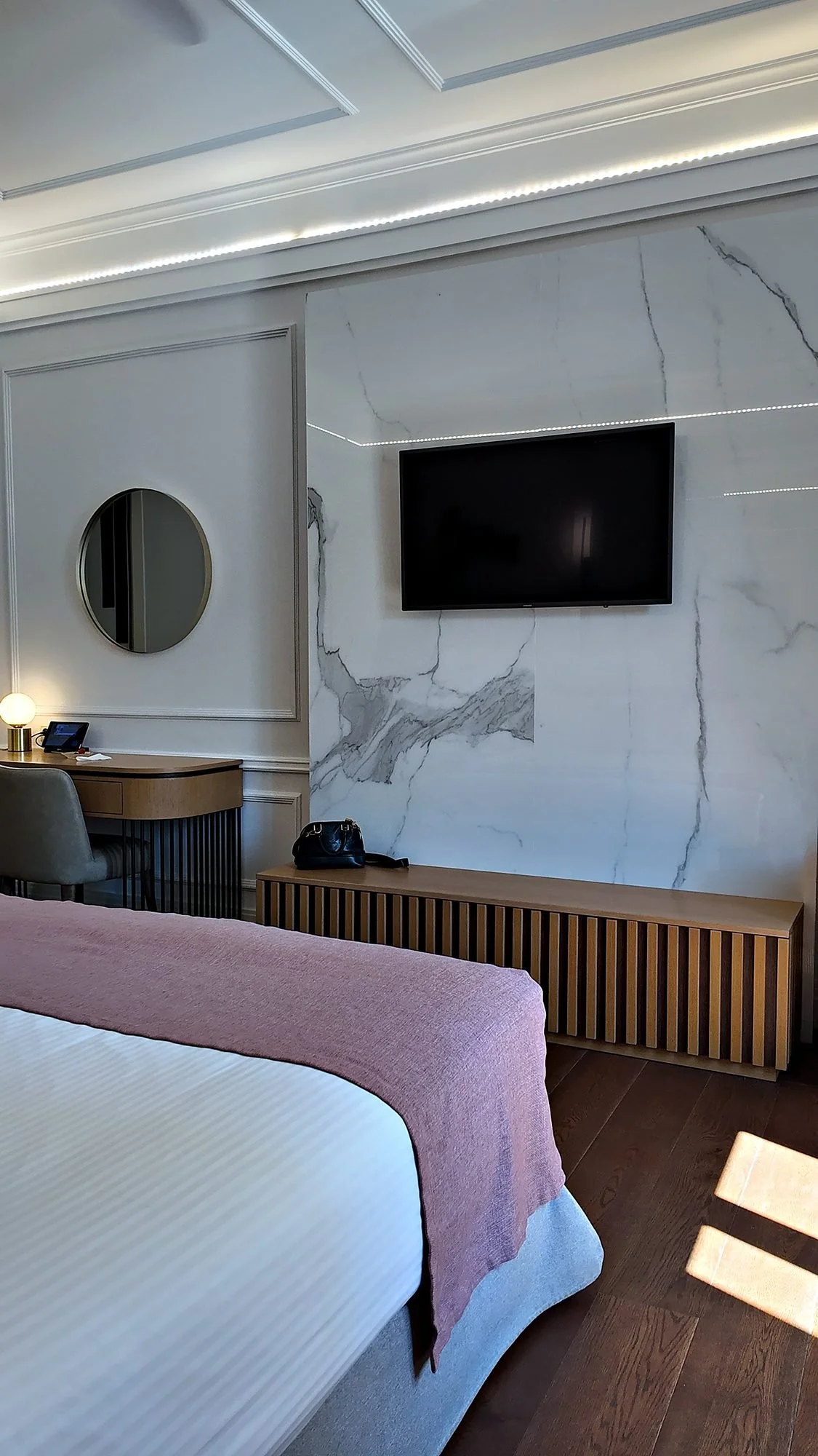 A marble feature wall, a small desk with a round mirror, and the end of the bed in an Elegant Suite at A77 Suites.