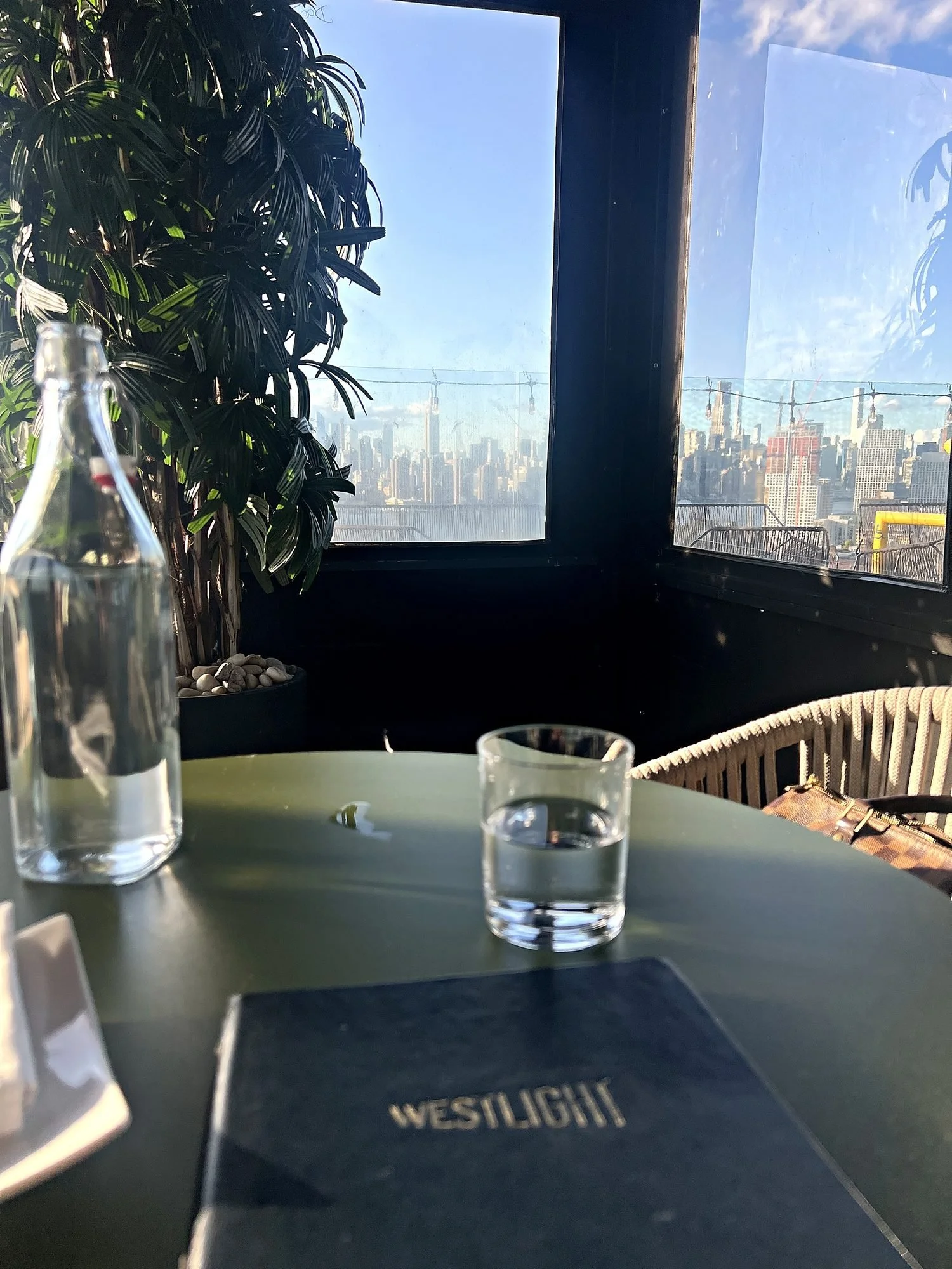 A corner table for one at Westlight, The William Vale's rooftop bar, with the menu on the table and the Manhattan skyline through the windows.