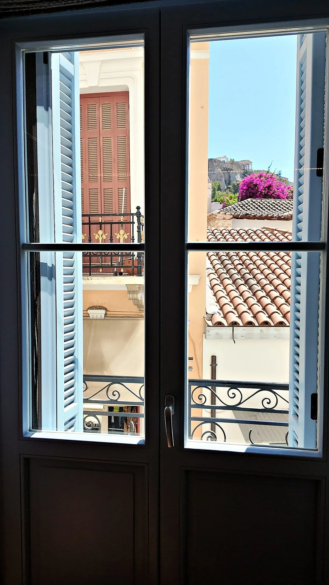 Tall French doors with blue shutters open onto a Juliet balcony at A77 Suites in Plaka, Athens, with the Acropolis beyond.