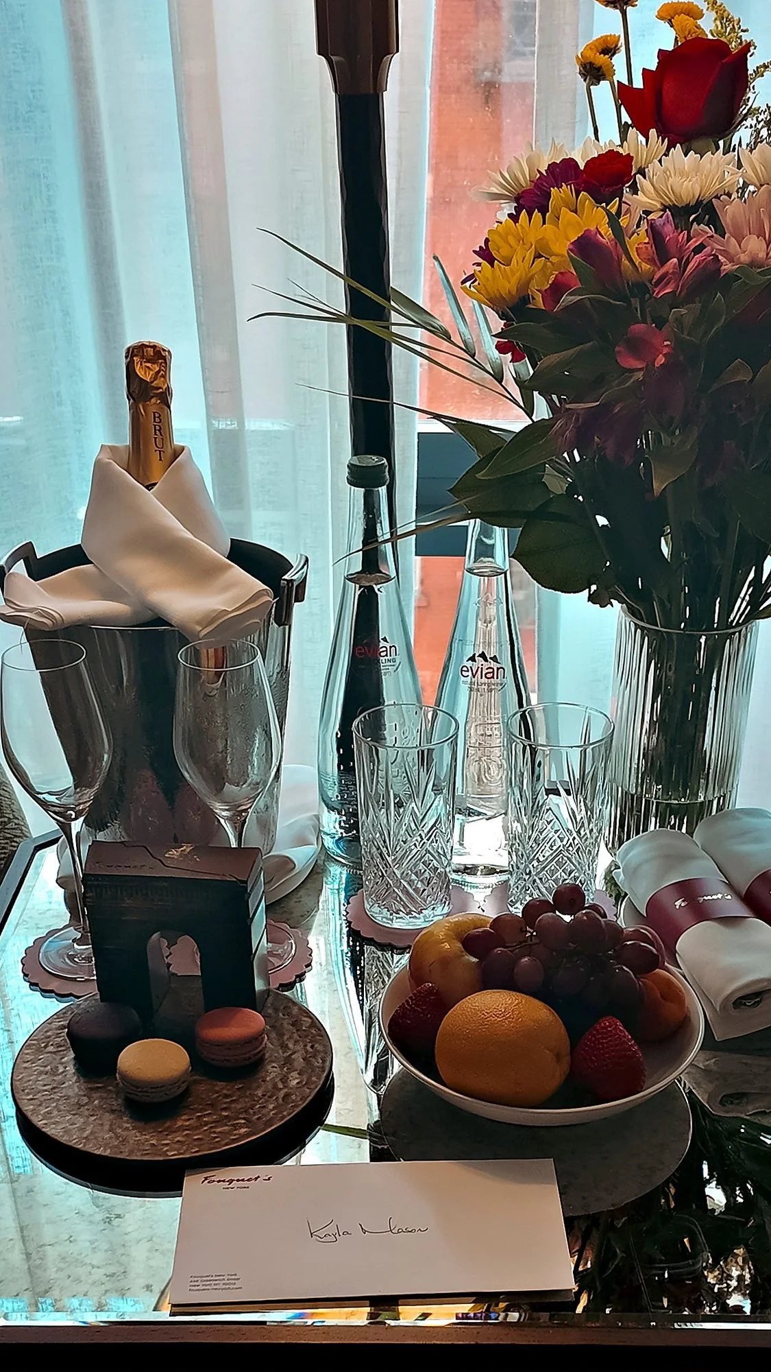 Flowers, champagne on ice, fruit and macarons on the welcome table in the room at Fouquet's New York, with a handwritten card in front.