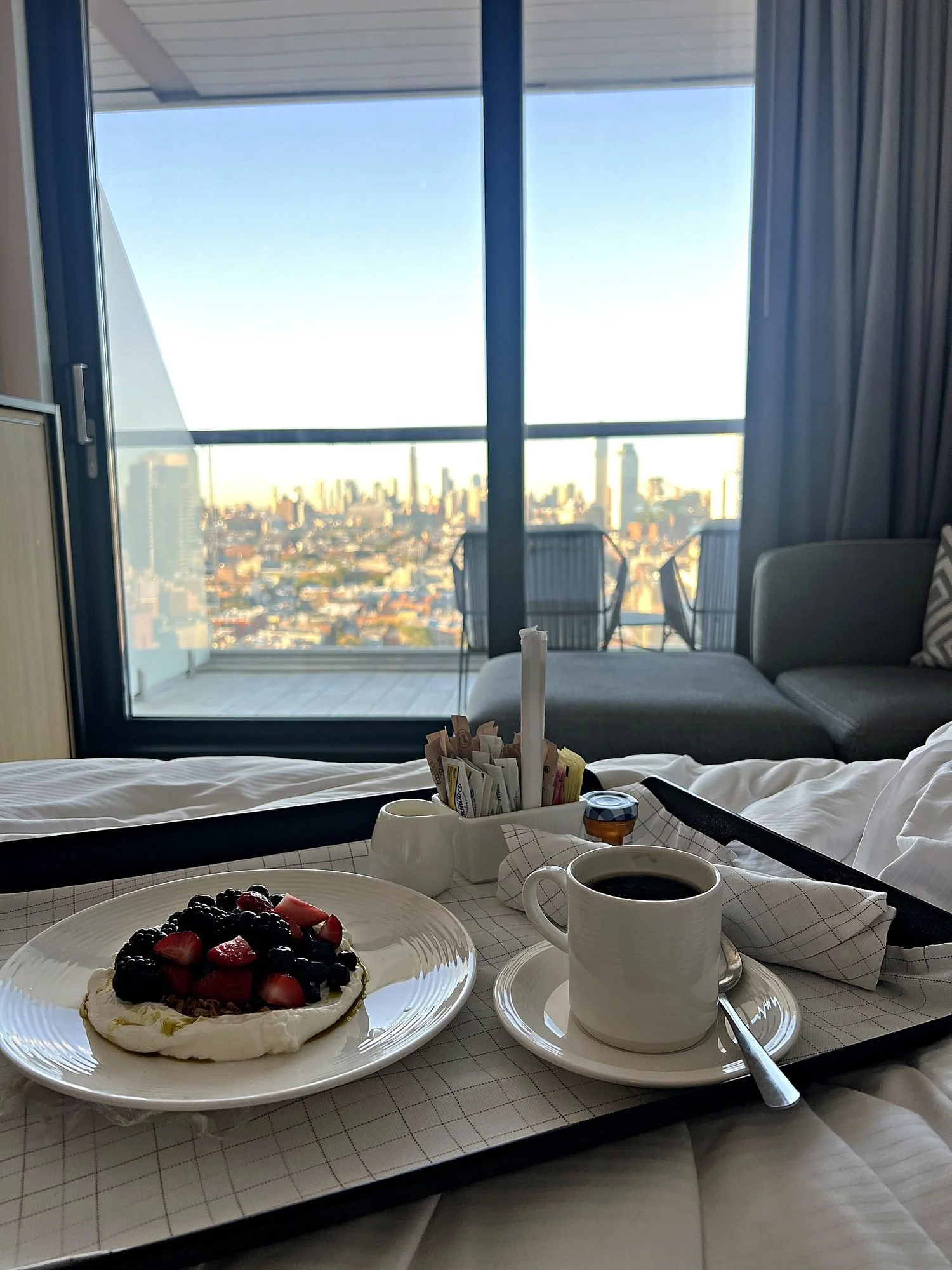 Breakfast on a tray in bed at The William Vale, yogurt with berries and coffee, with the balcony and the Manhattan skyline behind.