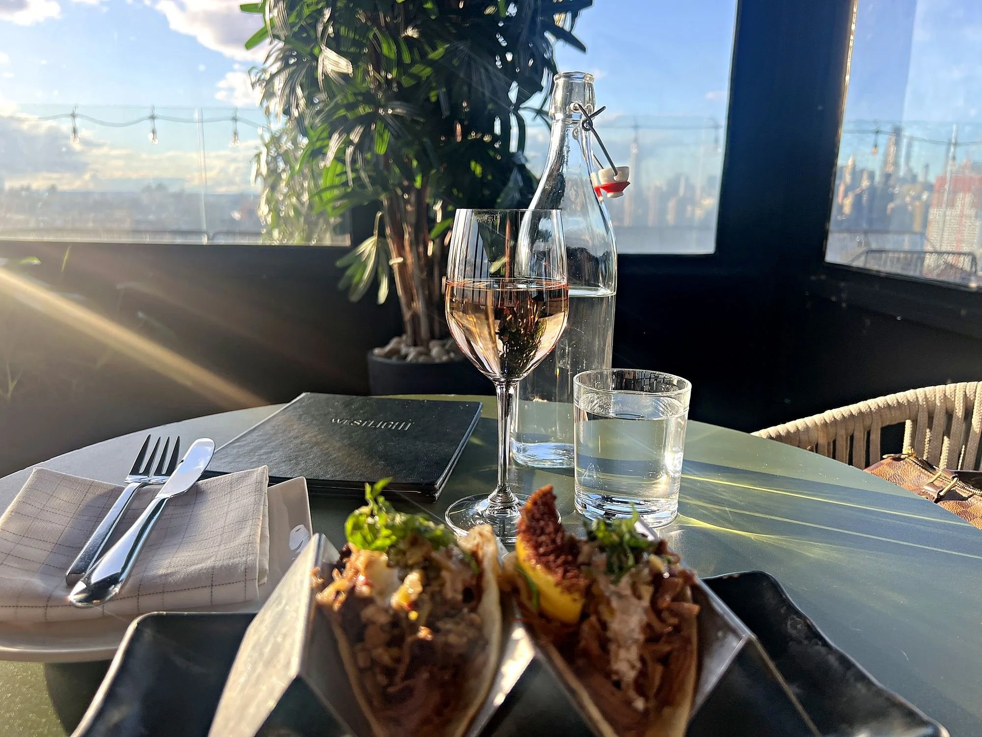 Shrimp tacos and a glass of rosé at a table for one at Westlight, the rooftop bar at The William Vale in Brooklyn, with Manhattan behind.