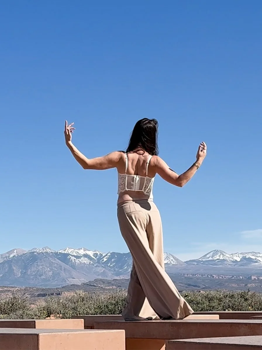 Woman in beige pants and a beige lace top dancing outdoors with snow-capped mountains in the background under a clear blue sky.
