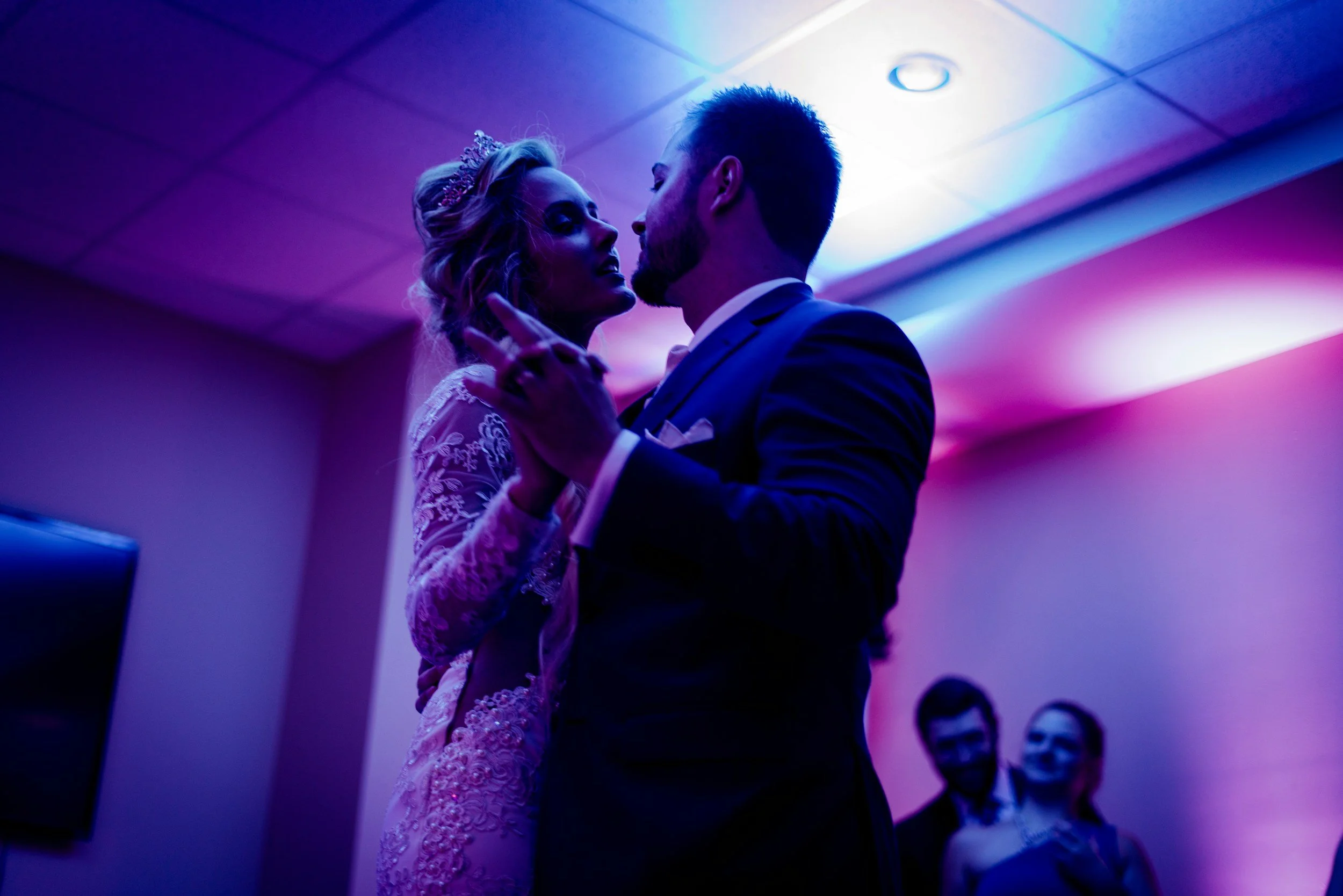 Couple dancing at a wedding reception, with purple and pink lighting and two people watching in the background.