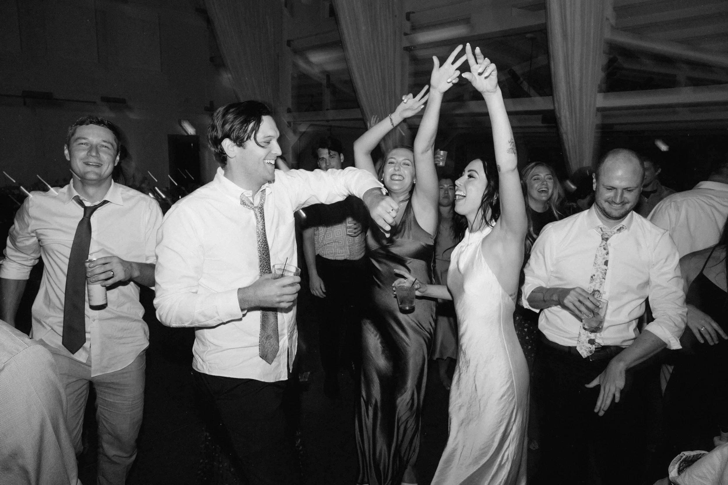 People dancing and celebrating at a party, with some holding drinks, in a dimly lit venue.