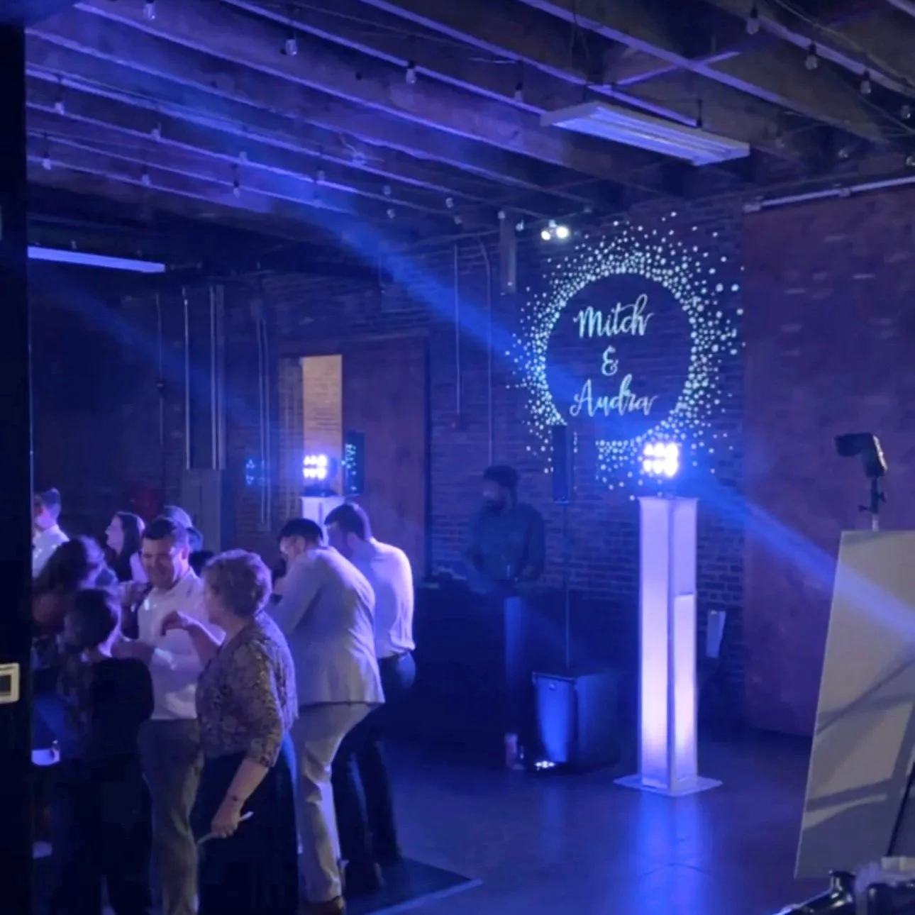 People gathered at a wedding reception with a decorated backdrop featuring the names Mitch and Audrey, illuminated by blue lighting. Gee Reed, DJ Gee6ix, Six Events, Photo Booths. monograms.