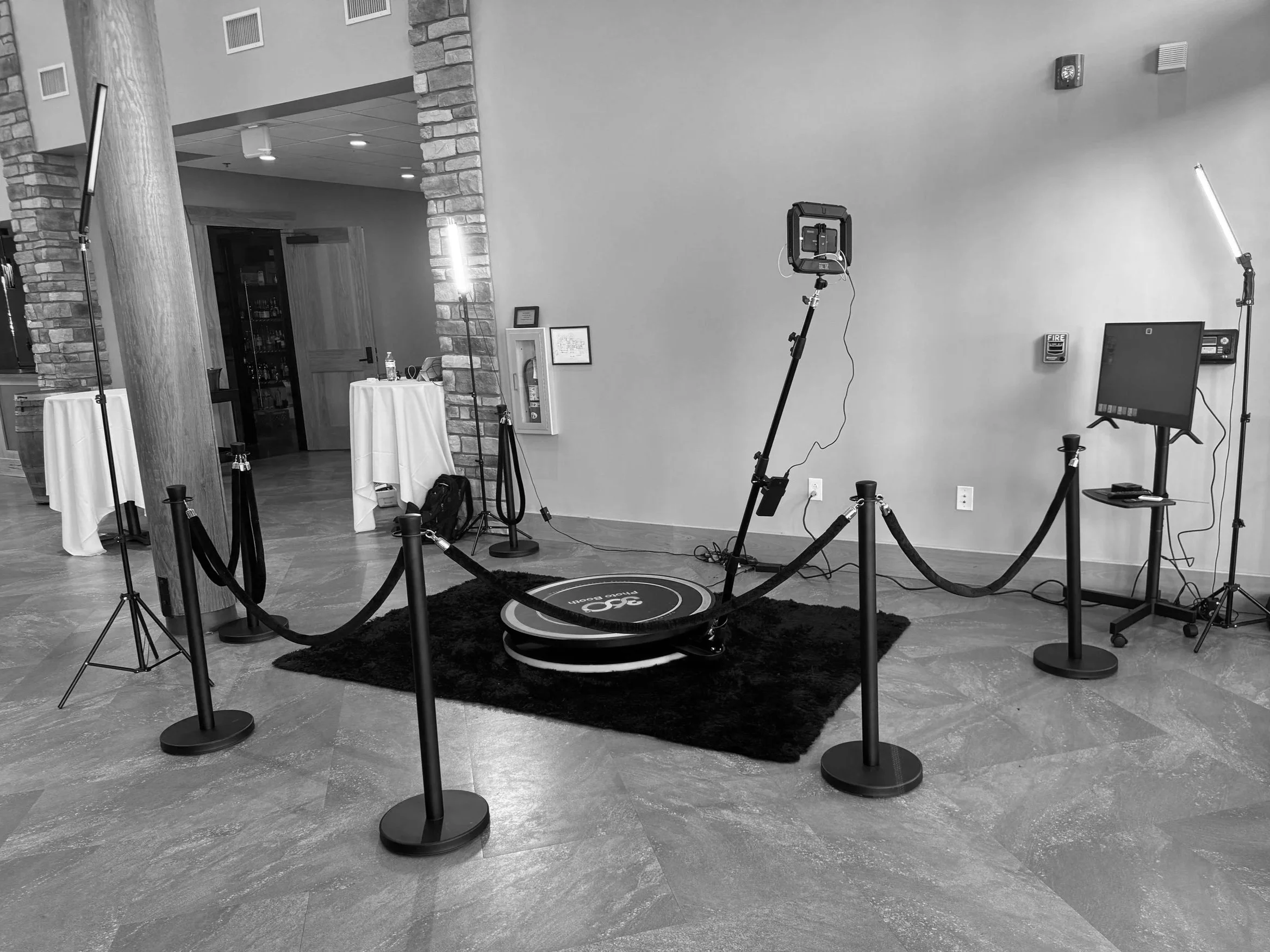 Set-up of 360 photo booth, surrounded by stanchions and ropes, in a room with tables, brick columns, and a wall with various equipment and decorations. Gee Reed, DJ Gee6ix, Six Events, Photo Booths, indy 360 booth,