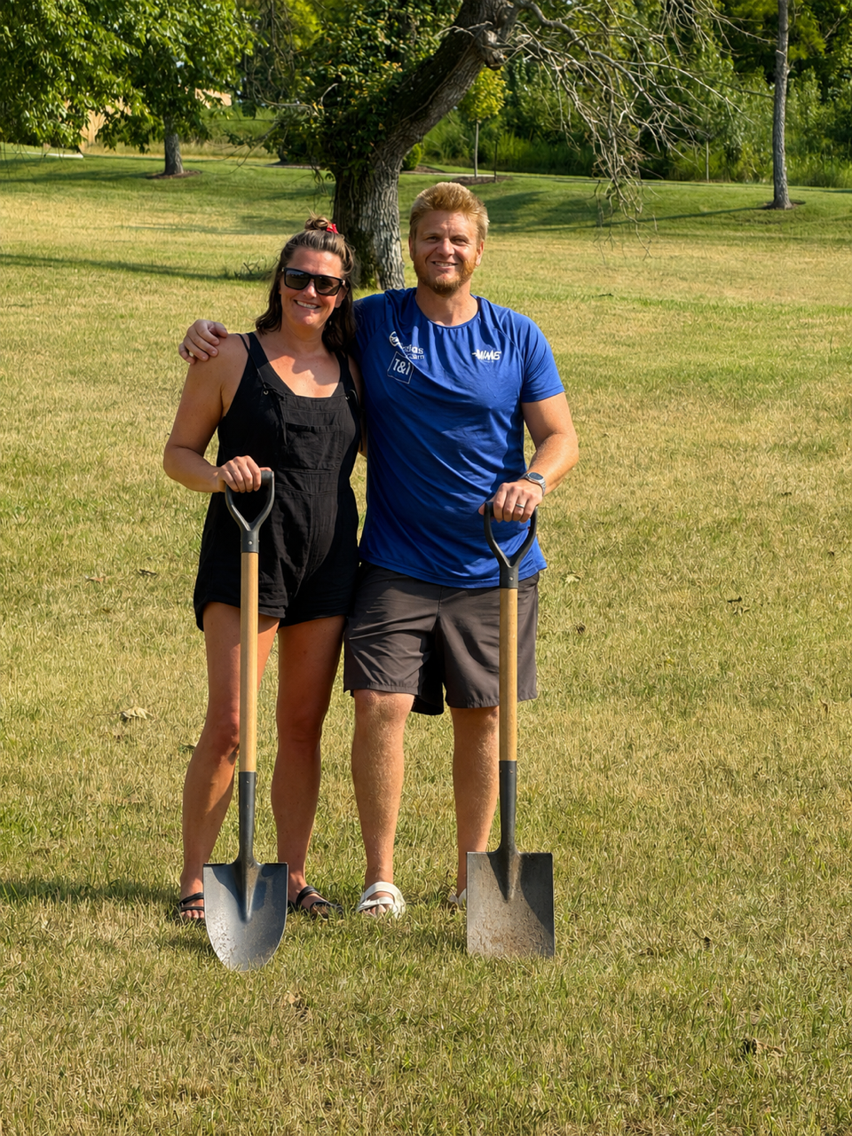 A man and woman standing outdoors on a grassy field, holding shovels, smiling, with a large tree in the background.