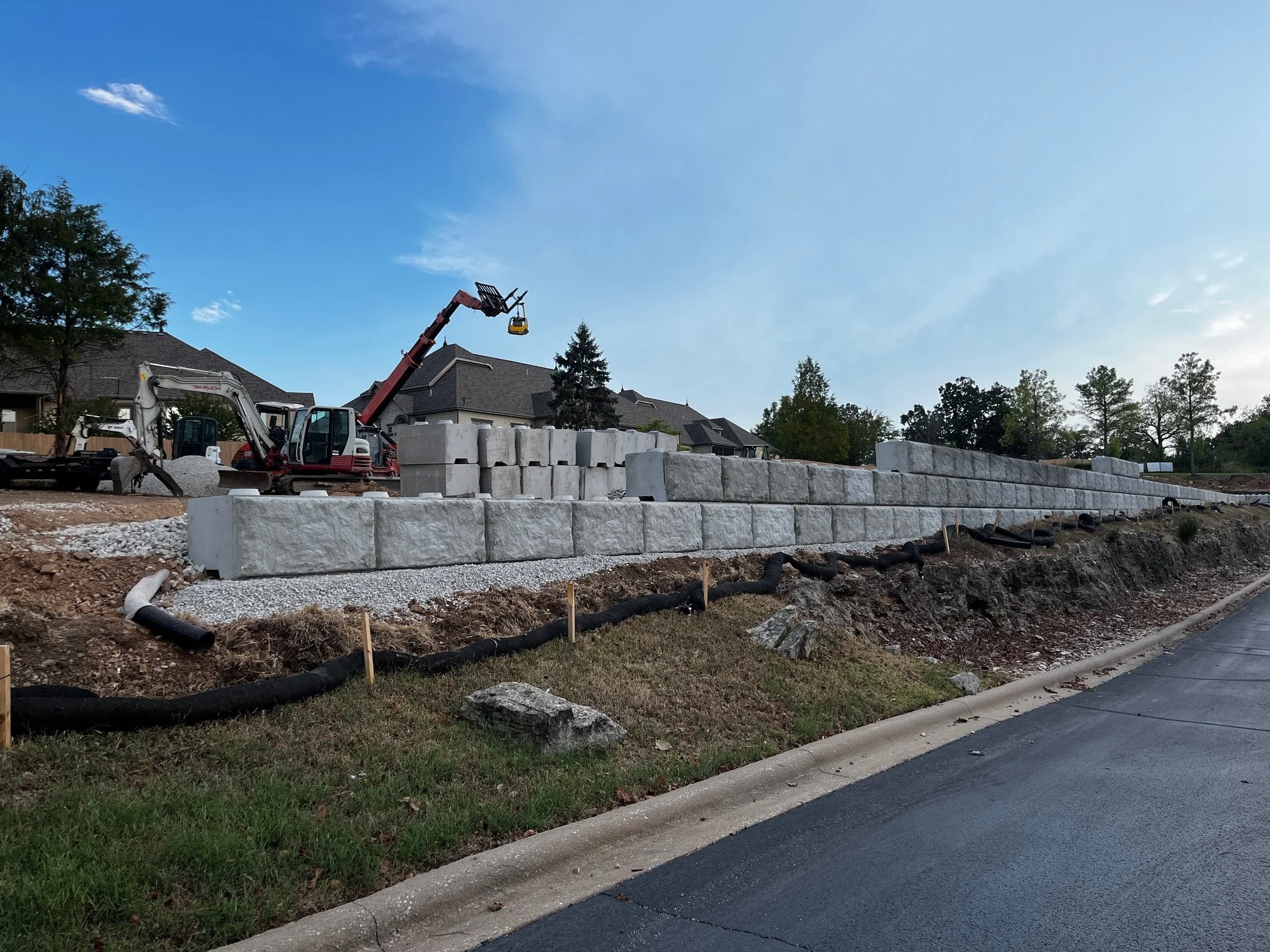Construction site with large stone blocks and machinery, houses in the background, and a clear blue sky.