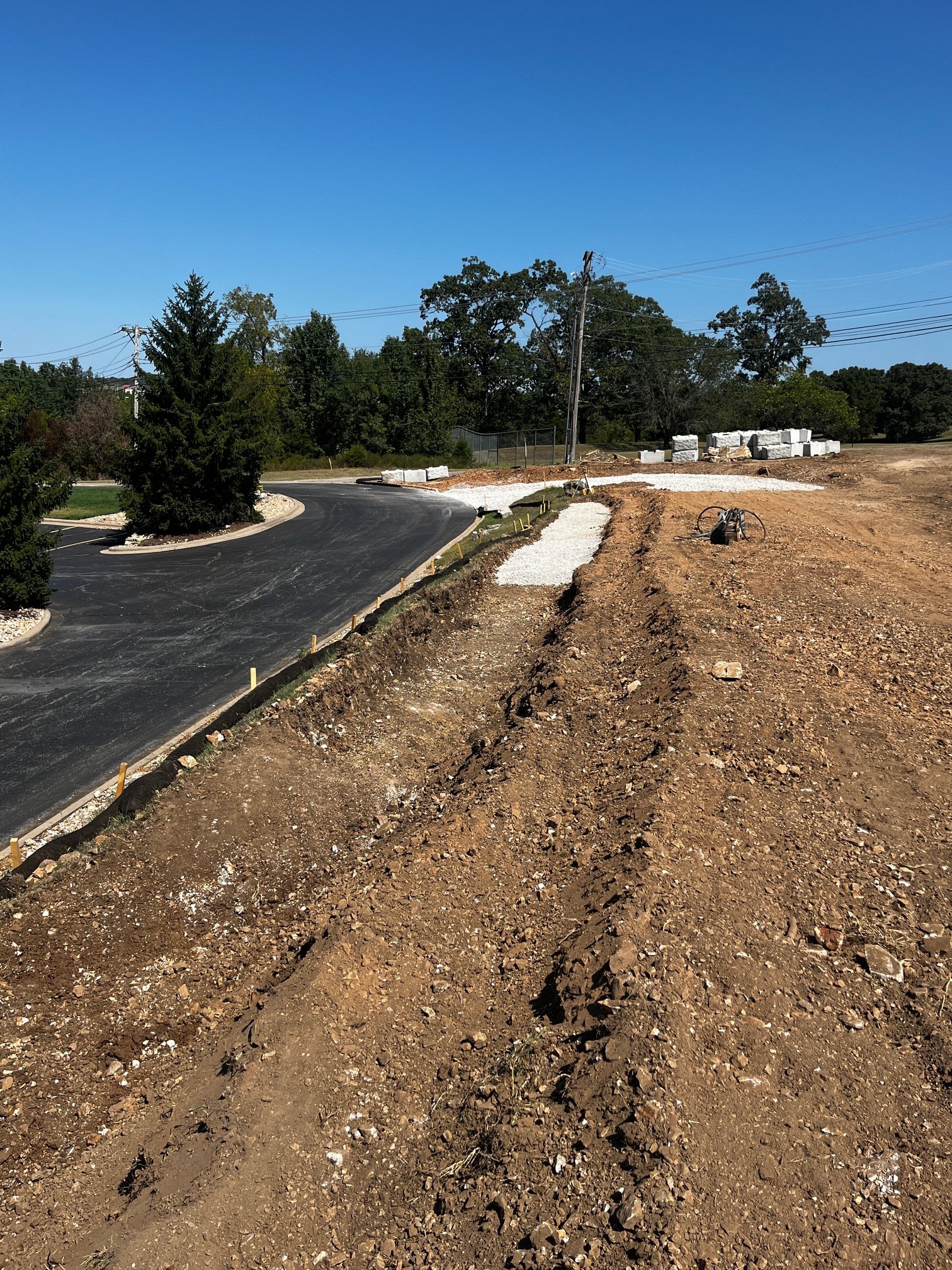Construction site with freshly paved road, dirt, and gravel, surrounded by trees and power lines under a clear blue sky.