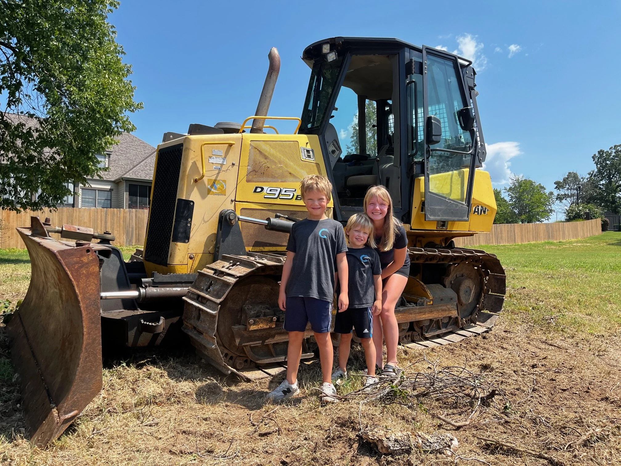 Three children and a woman standing in front of a large yellow and black bulldozer outdoors on a sunny day, with trees and a house in the background.