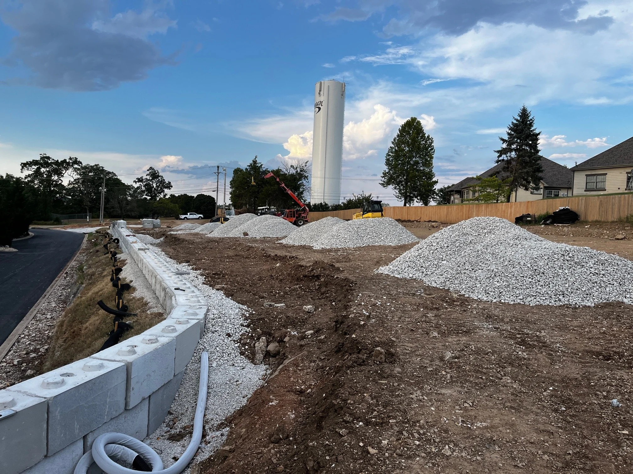 Construction site with gravel piles, construction machinery, a stacked wall of concrete blocks, and a partially built sidewalk, with residential houses and trees in the background under a partly cloudy sky.