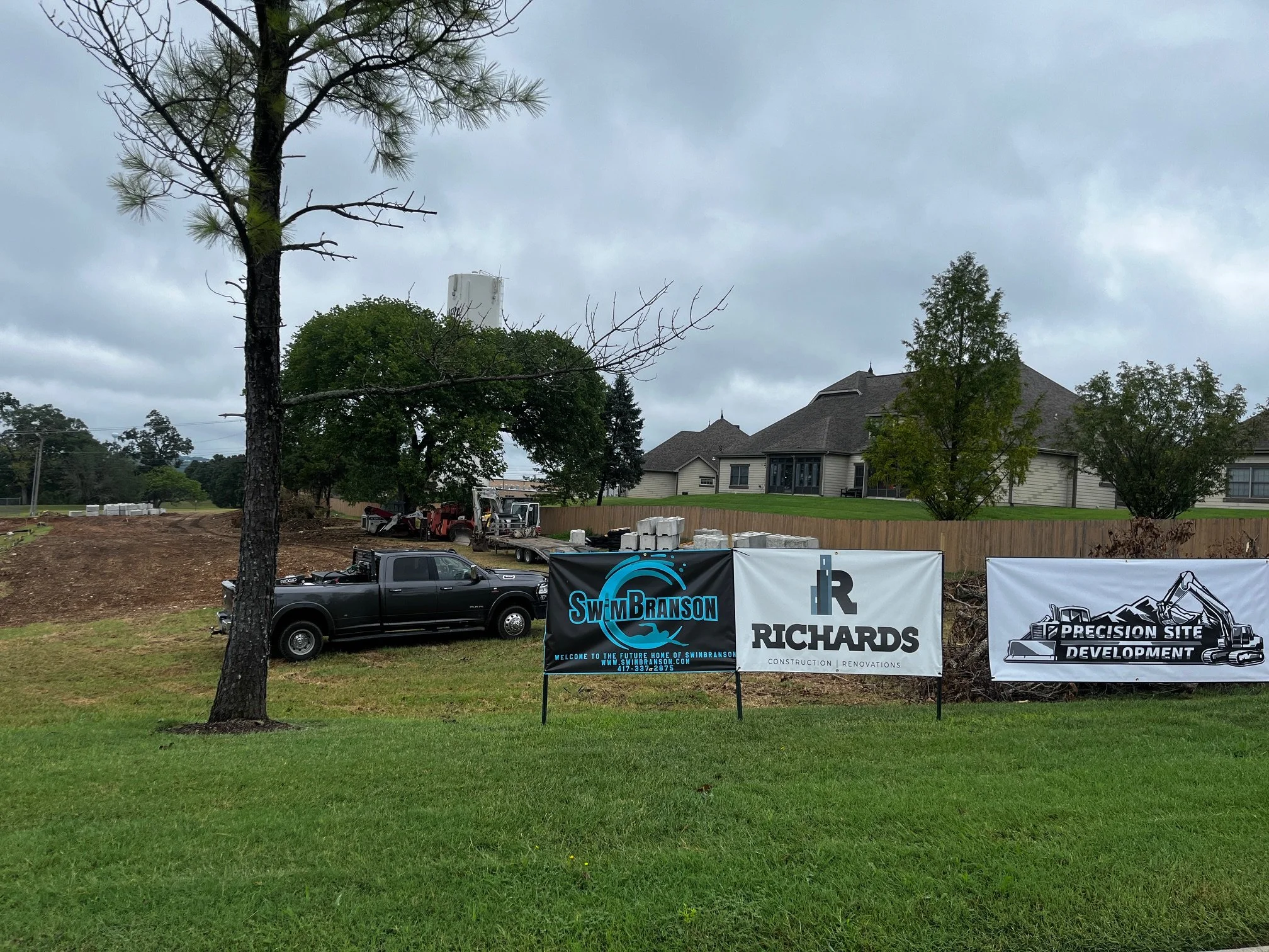 Construction site with banners for Swin Branson, Richards, and Precision Site Development, outdoor residential area with houses, trees, trucks, and cloudy sky.