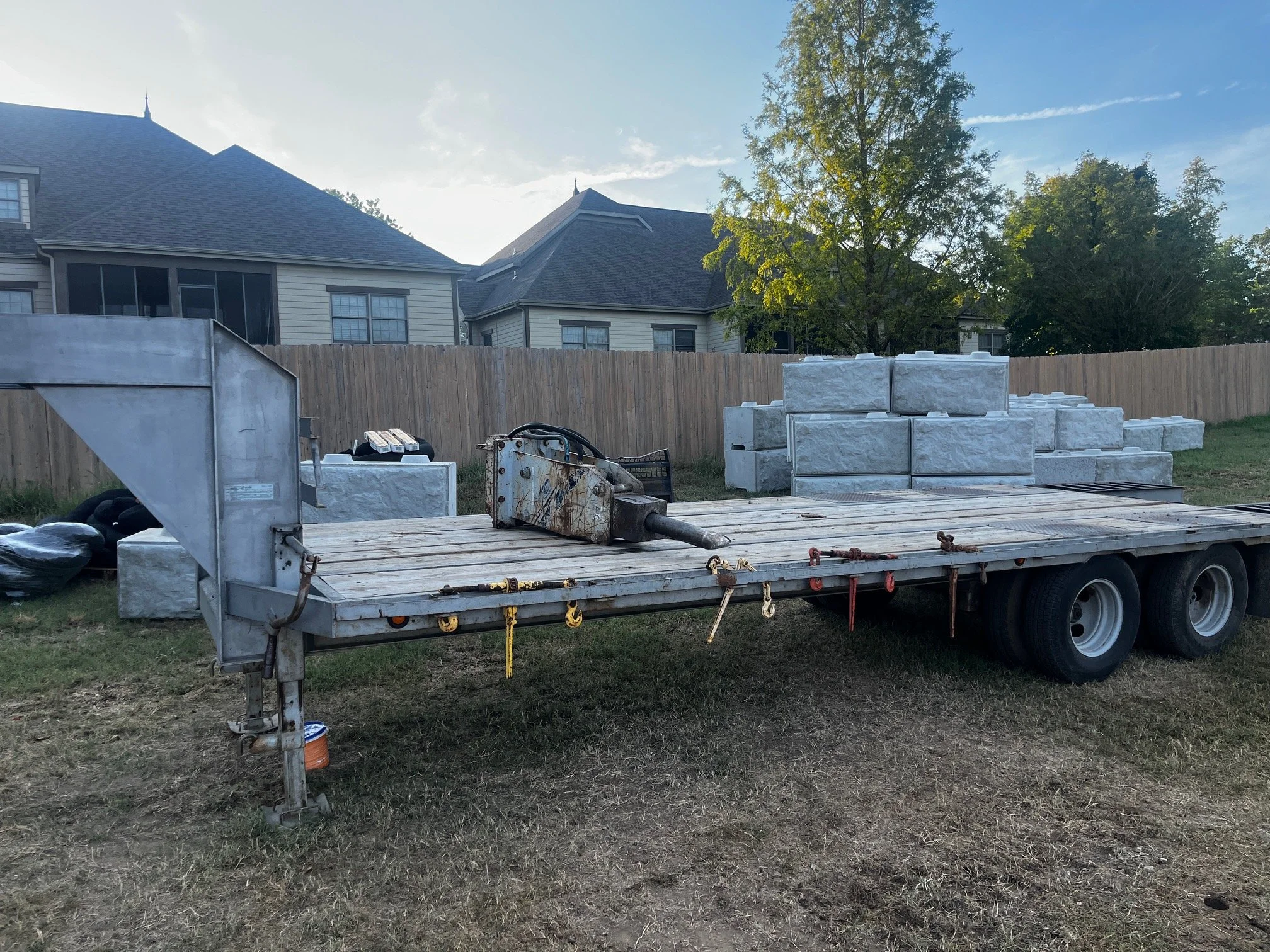 Construction site with a flatbed trailer loaded with stone bricks, a fence, and a house and trees in the background.