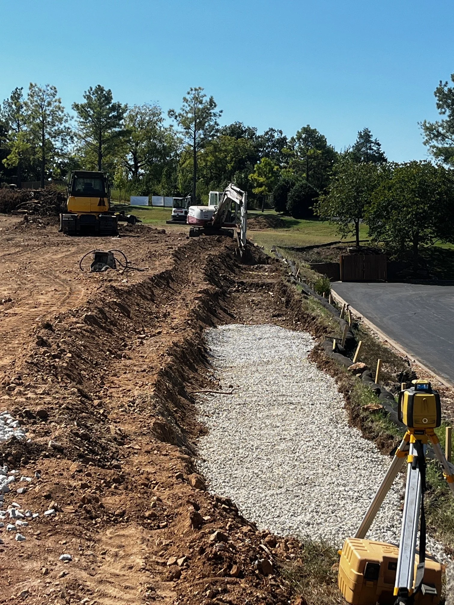 Construction site with an excavator, gravel pathway, surveying equipment, and trees in the background.