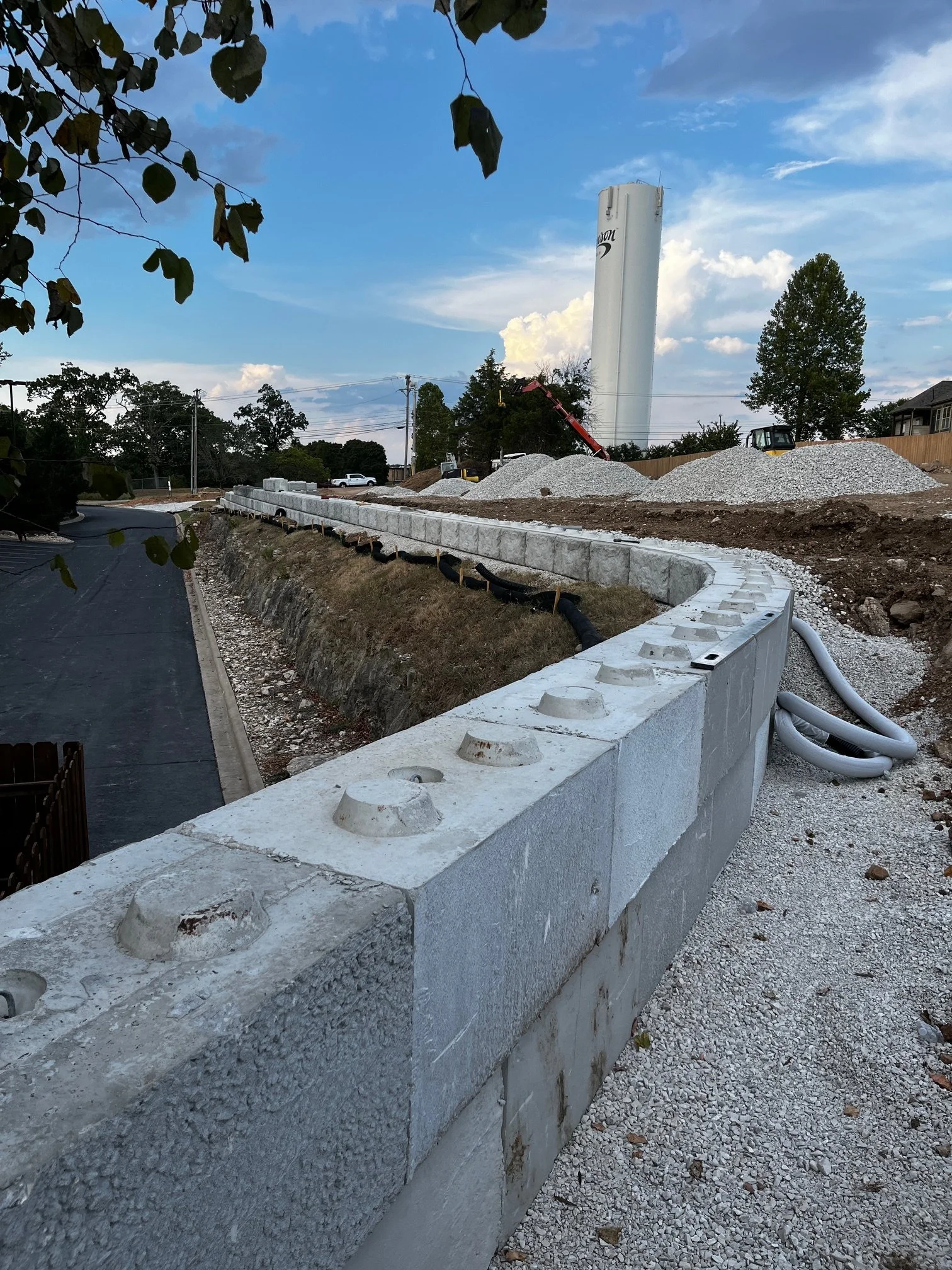 Construction site with a newly built concrete barrier, a pile of gravel, construction equipment, and a tall water tower in the background under a partly cloudy sky.