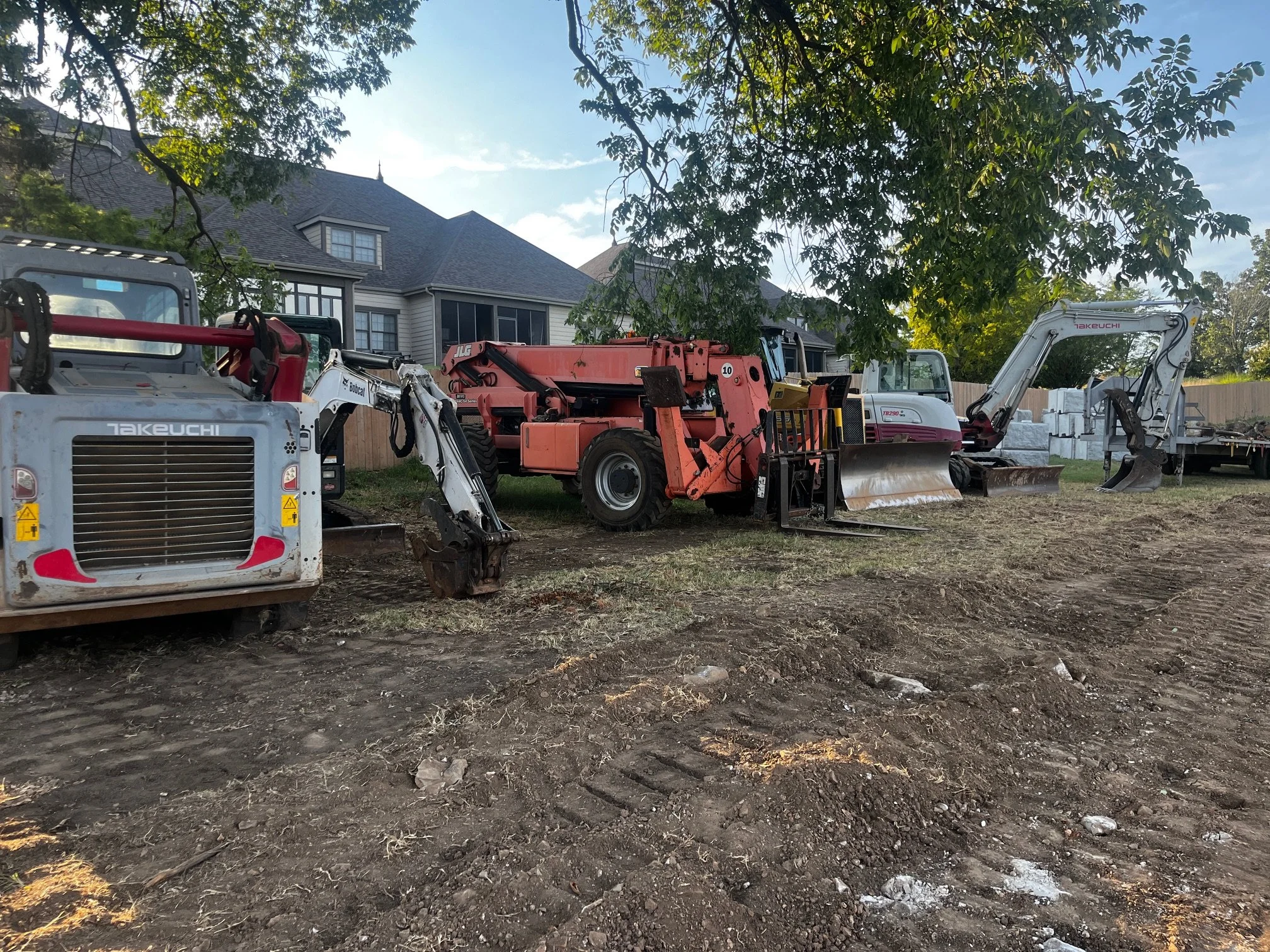 A backyard with construction equipment and machinery, including a small excavator and other tools, on a patch of dirt with a house and trees in the background.
