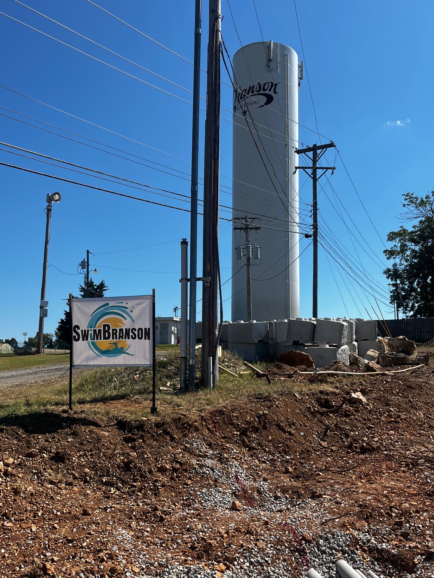A construction site with a large white water tower labeled 'Swim Branson' in the background, electrical poles and wires, a sign with the same logo, gravel and dirt ground, and a clear blue sky.