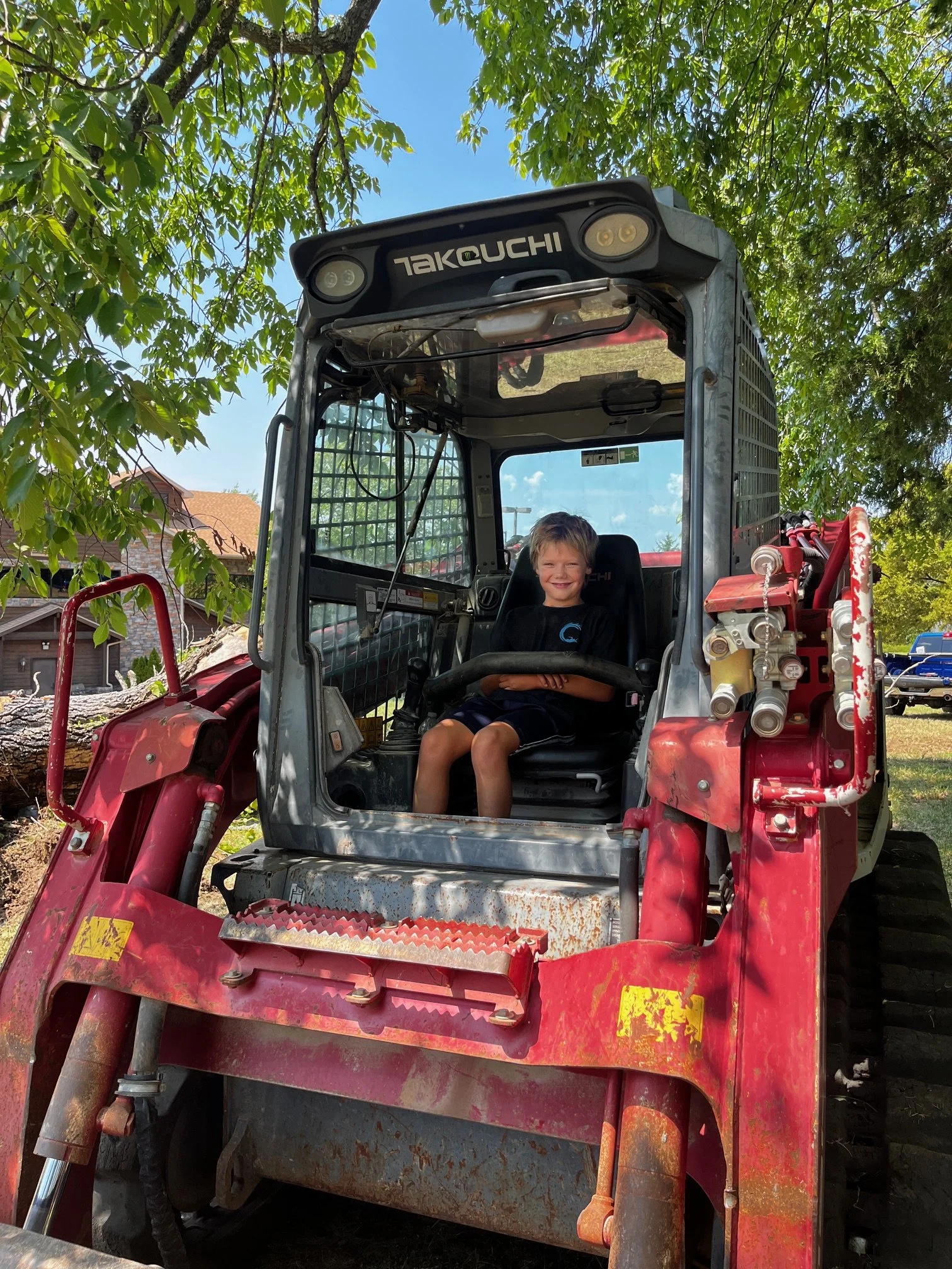 A young boy sitting in the driver's seat of a red tractor, outdoors under leafy green trees, with a house and parked cars in the background.