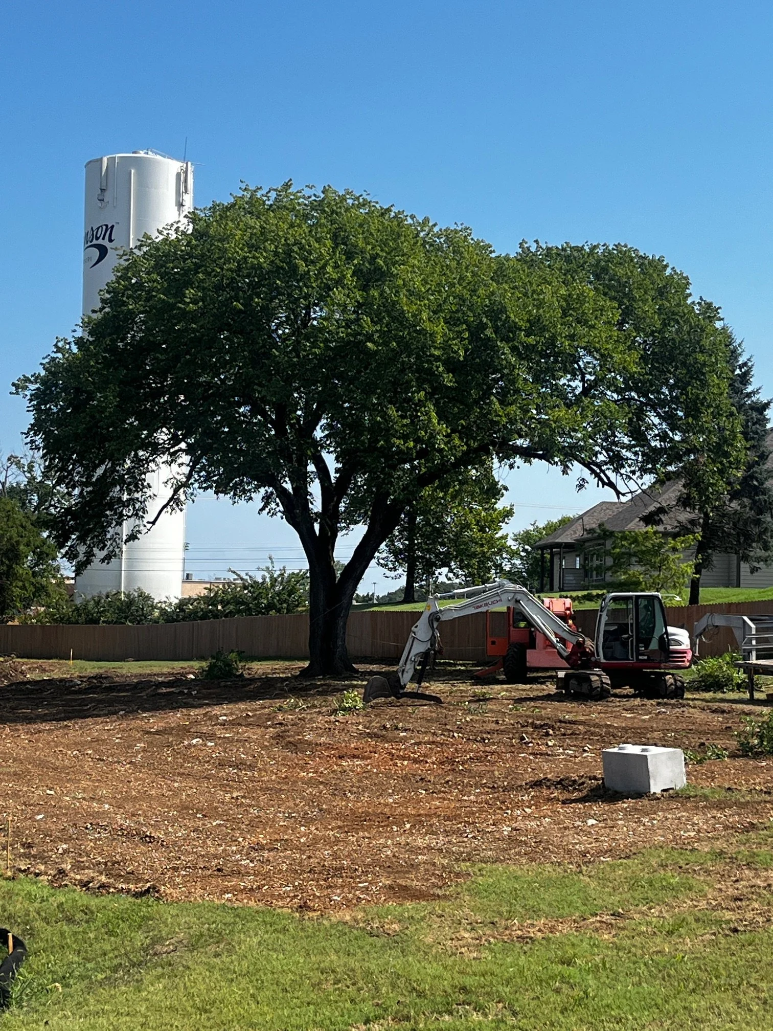 A construction site with trees, a small excavator, and a residential house in the background.