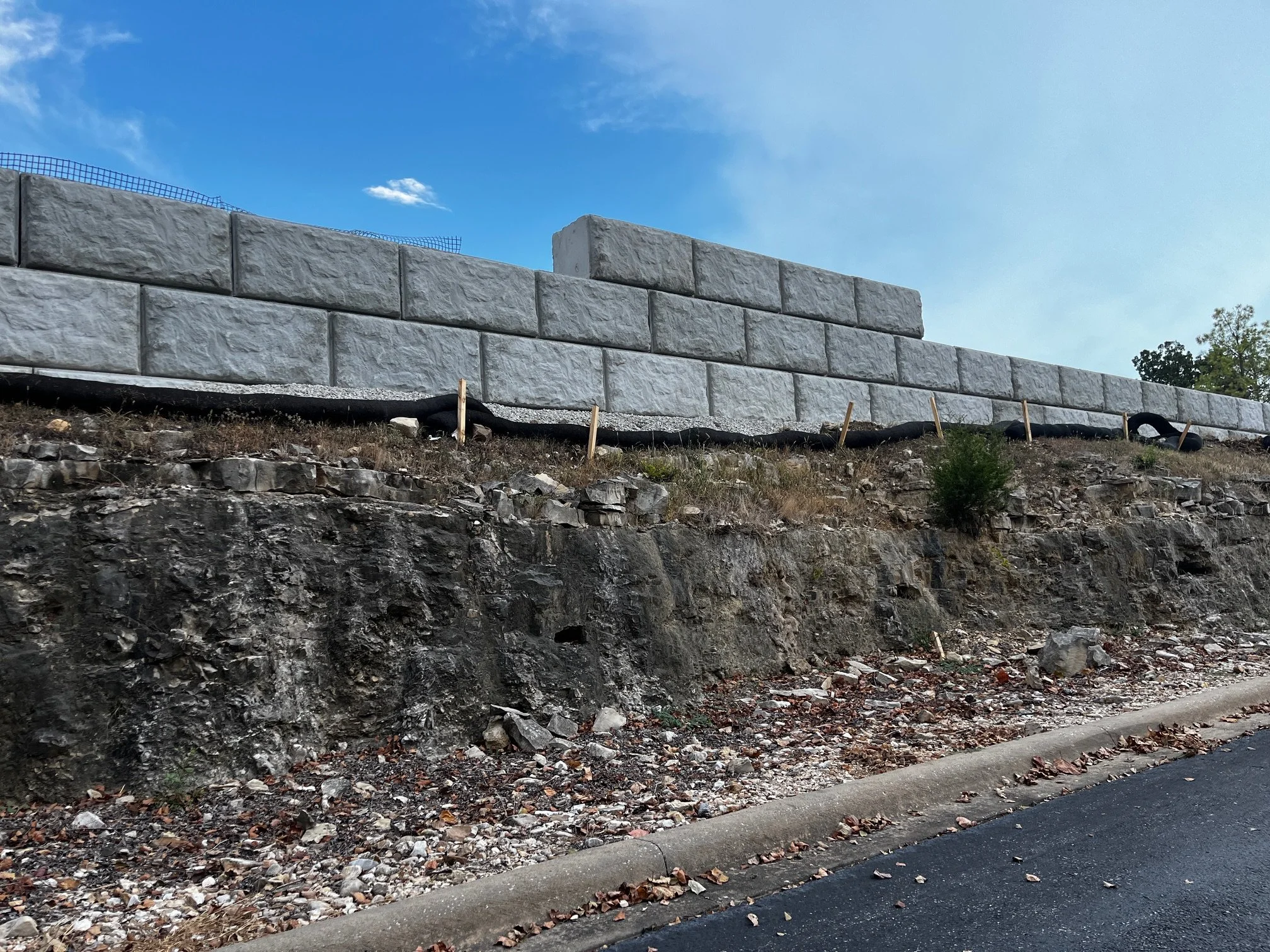 A retaining wall made of large concrete blocks on a slope with rocks and dirt, and trees in the background under a clear blue sky.