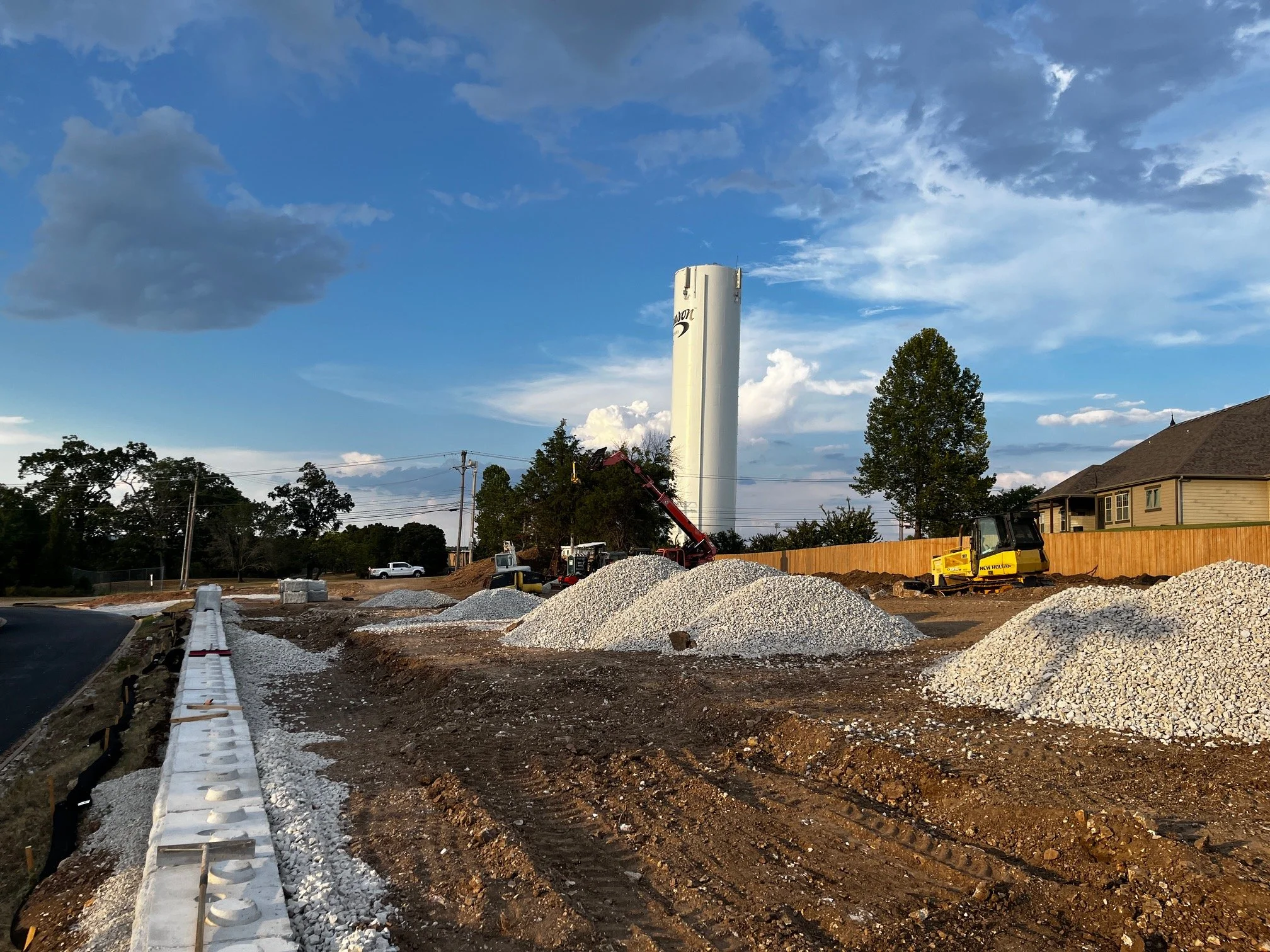 Construction site with gravel and white concrete blocks, a yellow and black excavator, and a tall white water tower against a blue sky with scattered clouds.