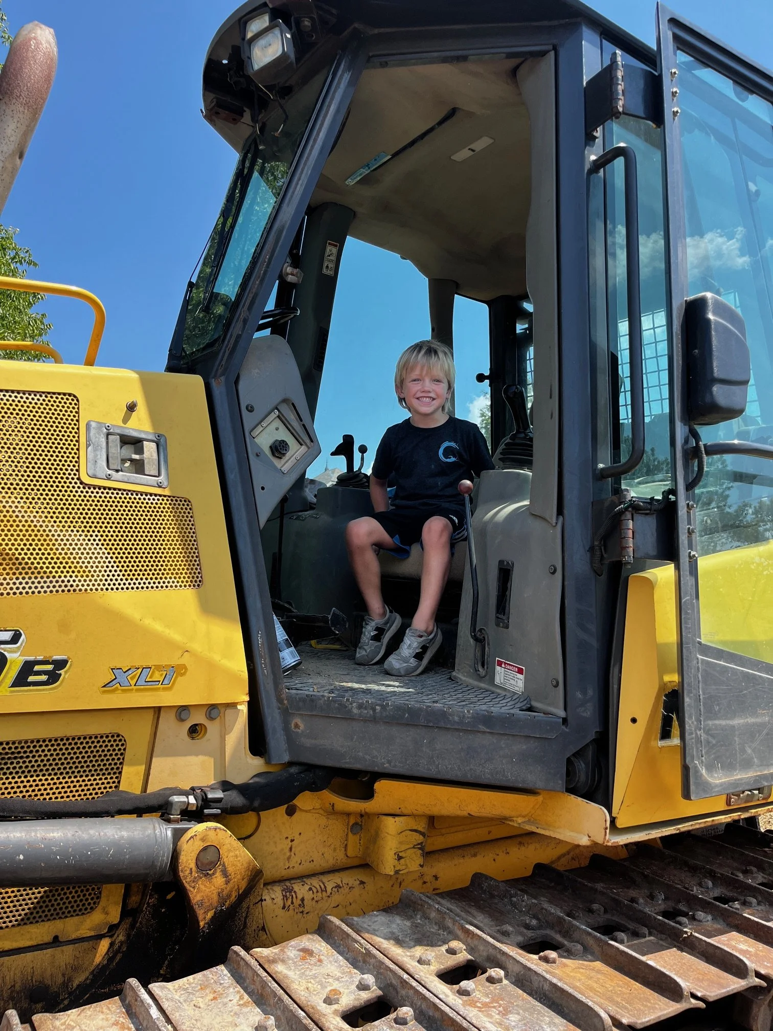 A young boy with blonde hair and a black shirt sitting inside the cab of a yellow bulldozer, smiling at the camera. The bulldozer has a large metal blade in the front and tracks on the ground, with a clear blue sky in the background.