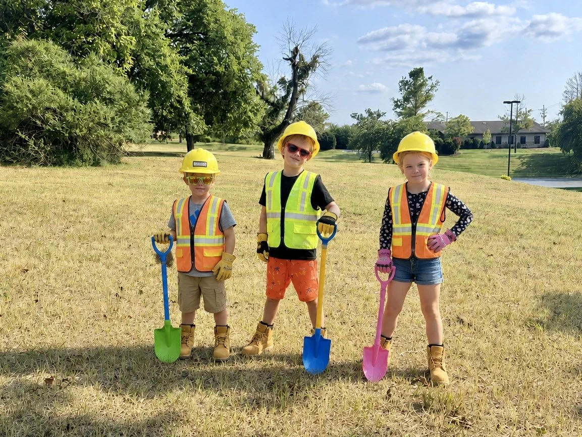 Three children wearing construction helmets, safety vests, and gloves, holding small shovels, standing on grass in a park-like setting on a sunny day.