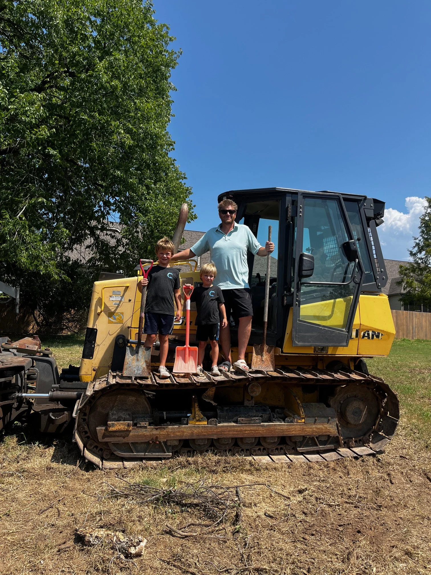 A man and two children standing on a yellow bulldozer in a backyard, with a tree and a house in the background, under a clear blue sky.