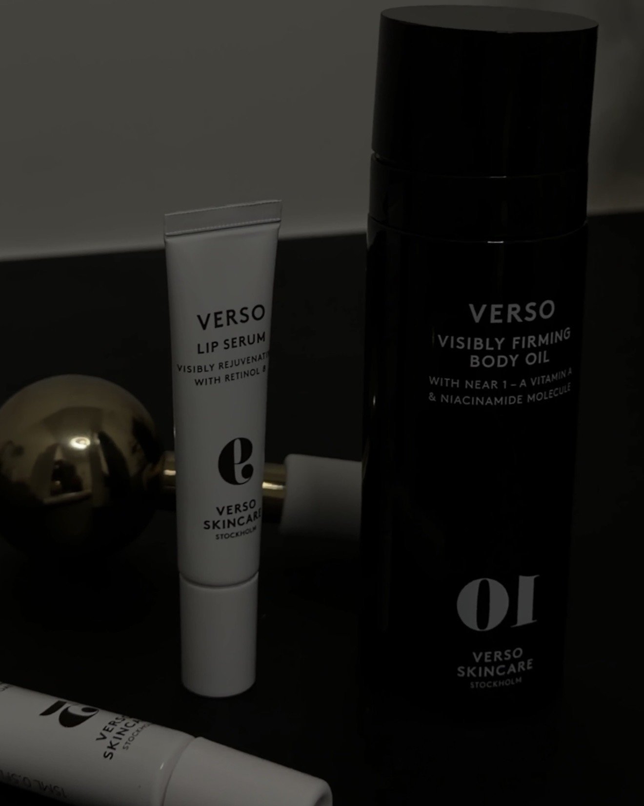 A black countertop with skincare and body products, including a white tube labeled 'Verso Lip Serum' and a black bottle labeled 'Verso Visibly Firming Body Oil', along with a gold decorative sphere and white and gold boxes.