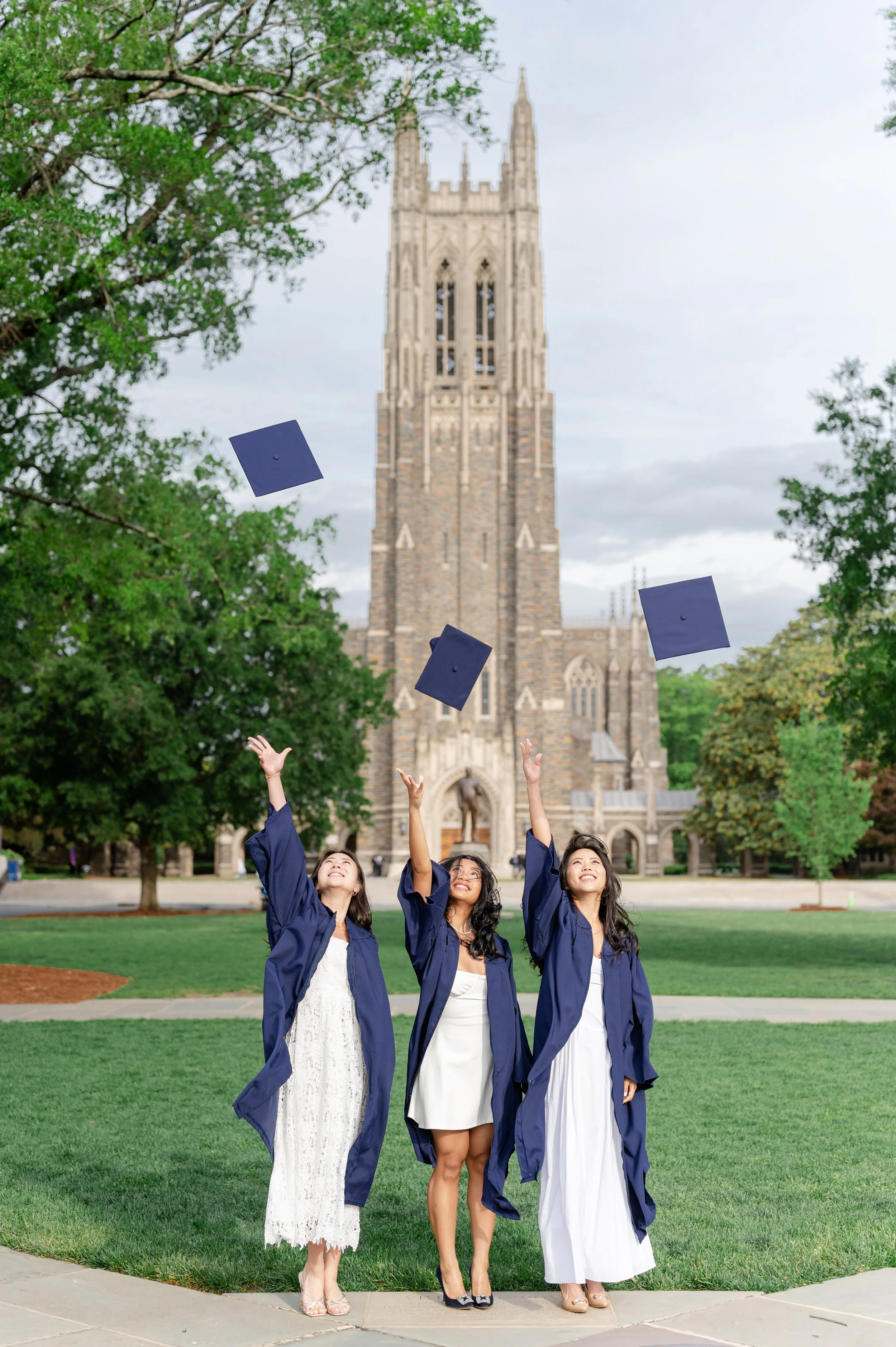 Three women in graduation gowns tossing hats in the air on a university campus with a large stone church or cathedral in the background.
