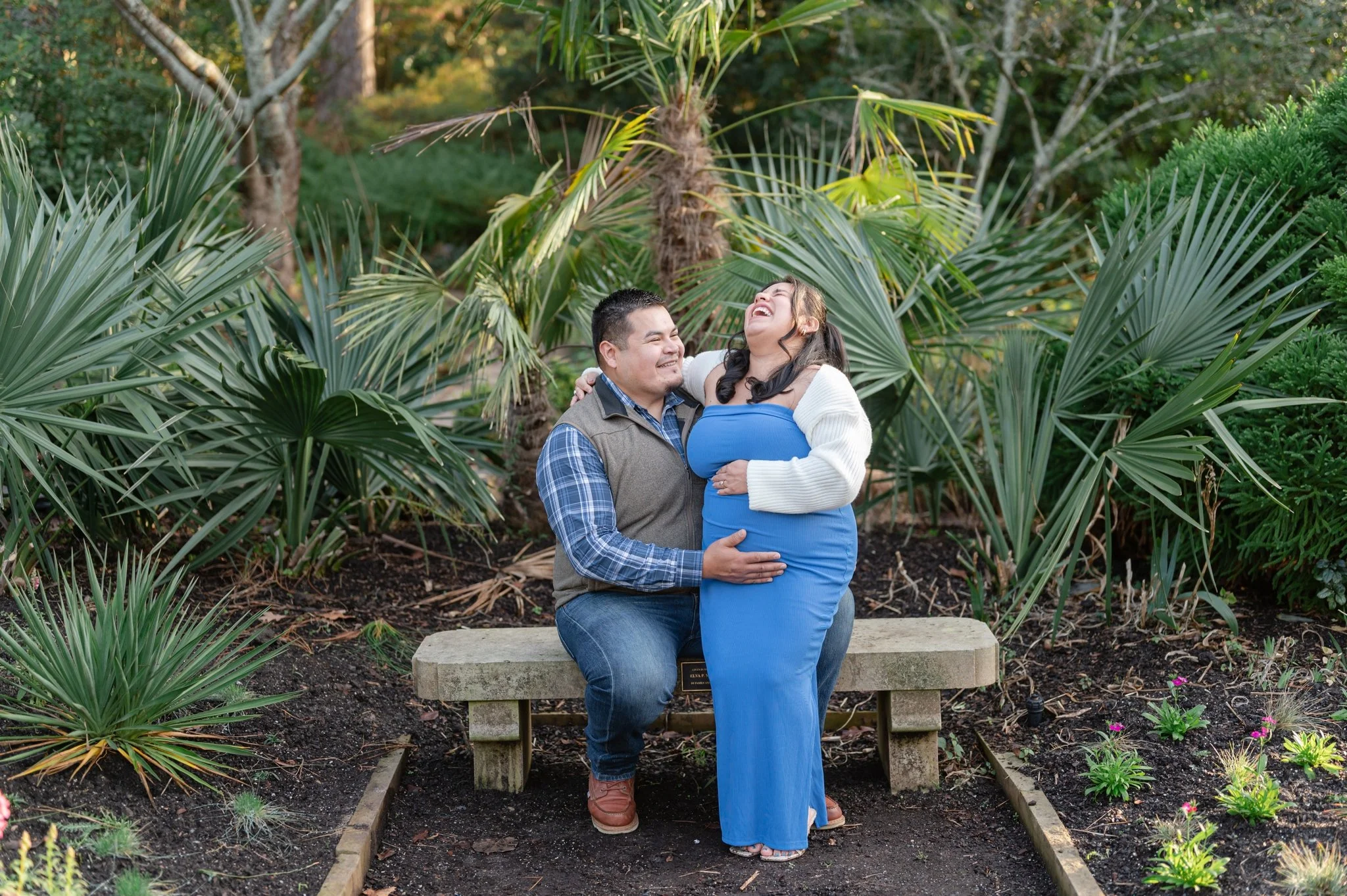 A latino couple laughing and embracing each other on a park bench in a lush, green garden with various plants and trees.