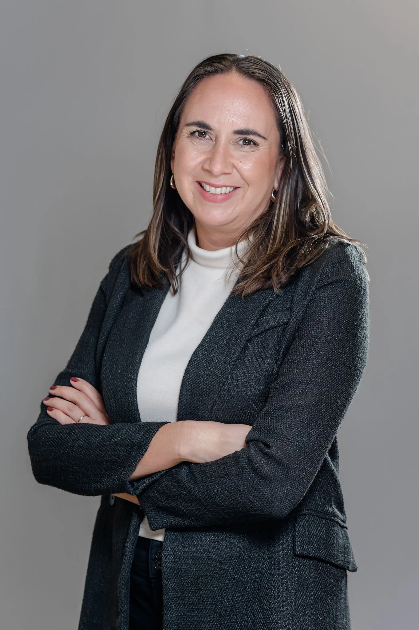 A woman with brown hair wearing a white turtleneck and a dark blazer, smiling and crossing her arms against a gray background.