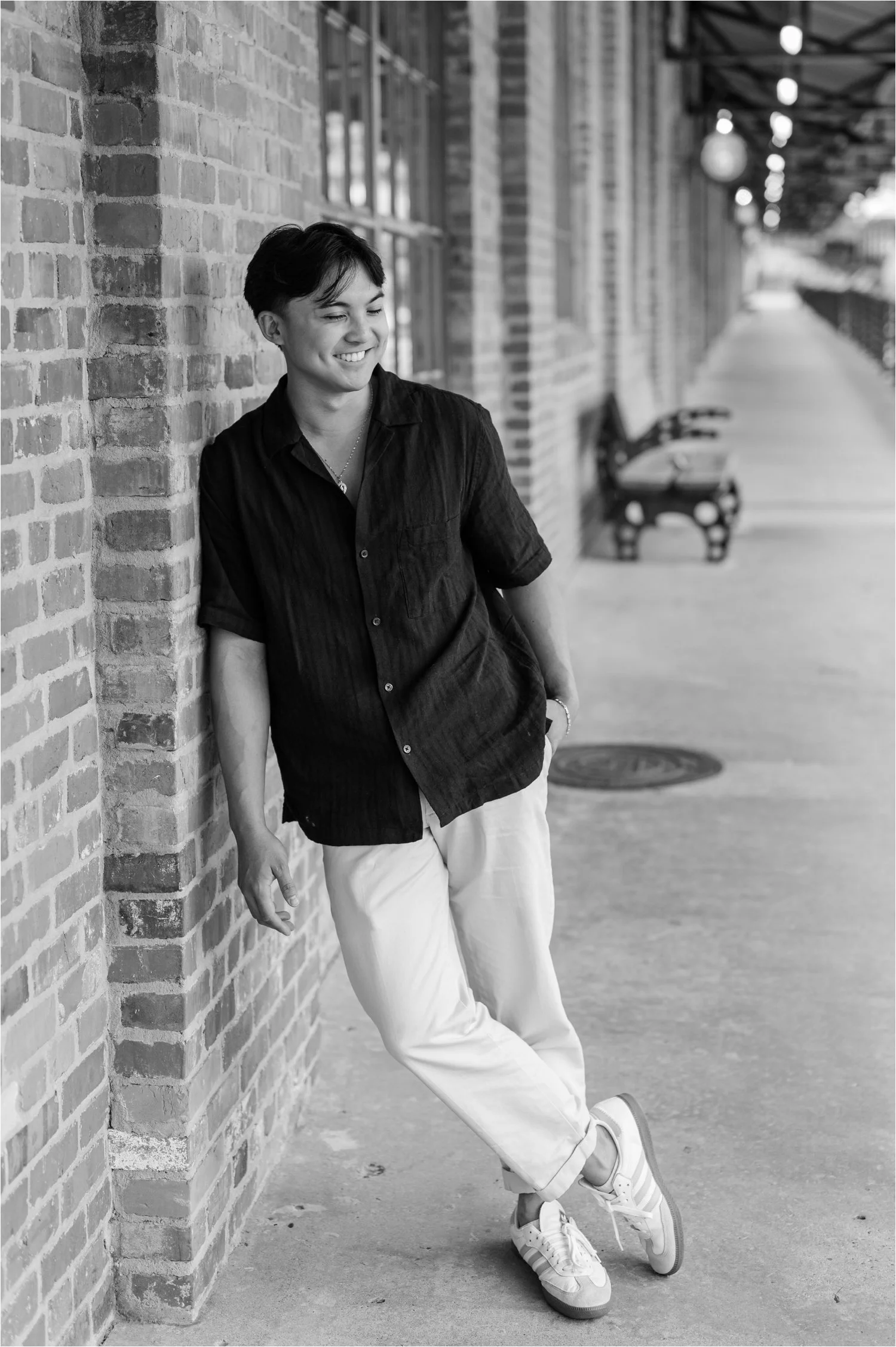 Young man with short dark hair smiling, leaning against a brick wall on a porch, wearing a dark shirt, white pants, and sneakers, with benches and hanging lights visible in the background.